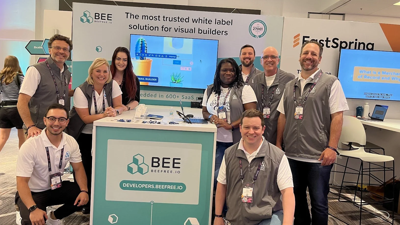 Group of nine smiling people at a BEE conference booth, with a large screen displaying the BEE logo and a monitor showing a presentation. The booth features BEE branding and a sign that reads 'Developers.beeFREE.io'.