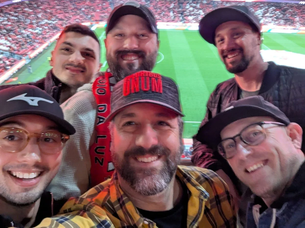 Six men smiling at a sports stadium, taking a group selfie with the field in the background.