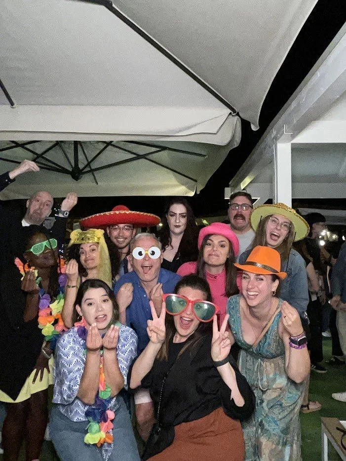 Group of people wearing colorful and playful costumes, including hats, large glasses, leis, and accessories, at a lively party or celebration outdoors at night.