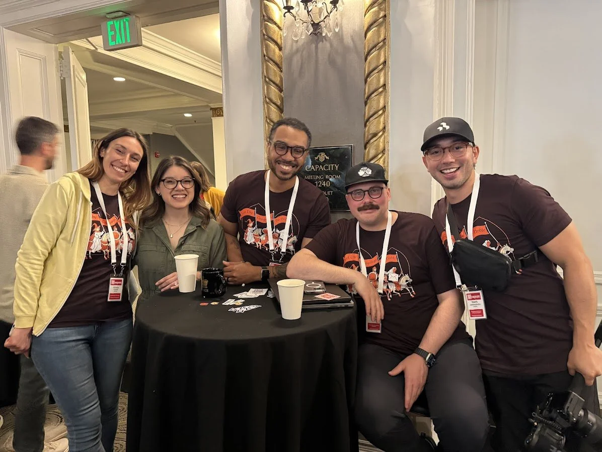 Group of five people smiling and posing together around a table at an indoor event or conference, with some wearing matching t-shirts and lanyards.