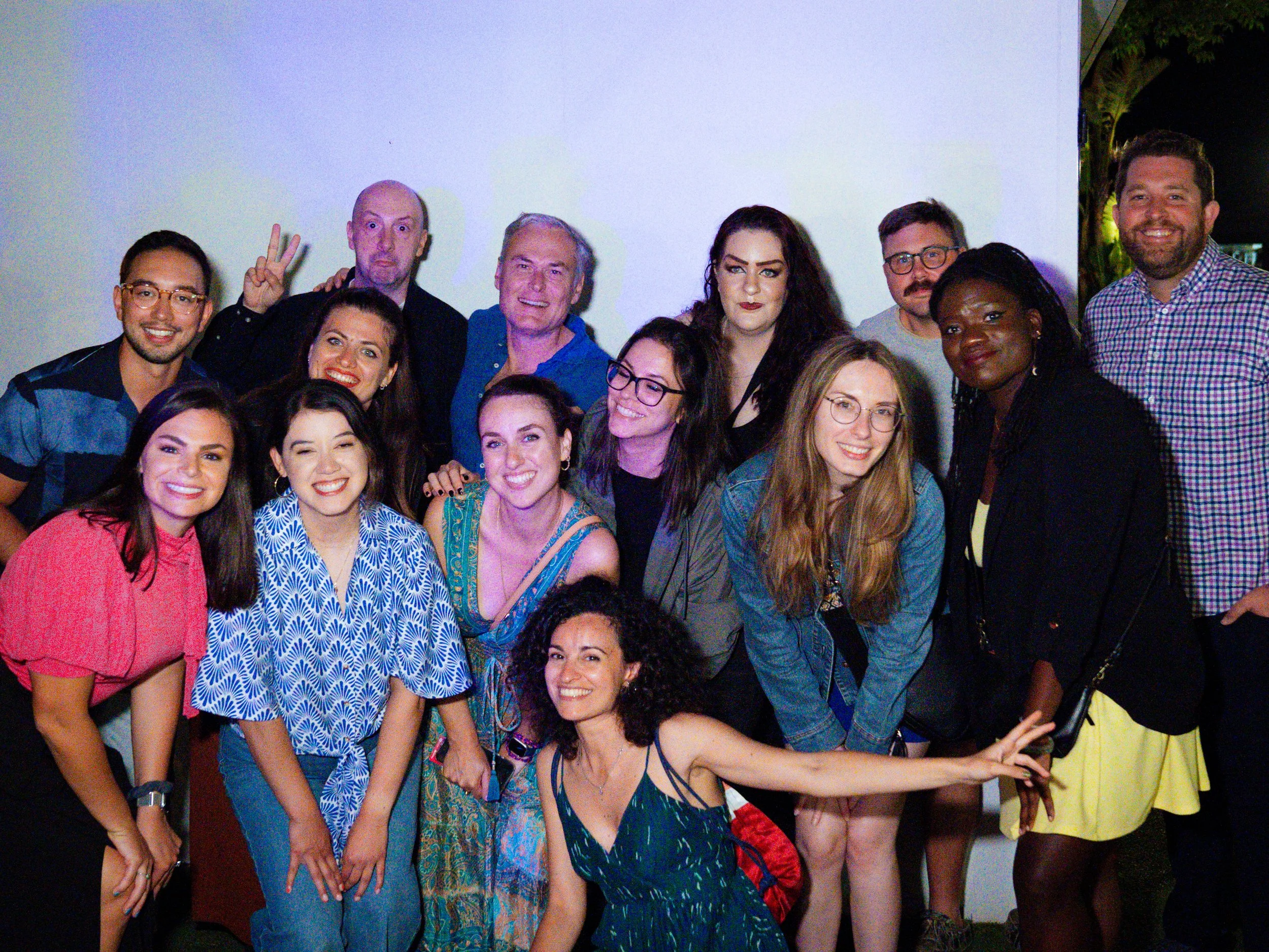 A group of diverse people posing together at a social event, standing in front of a plain white wall, smiling, and making playful gestures.