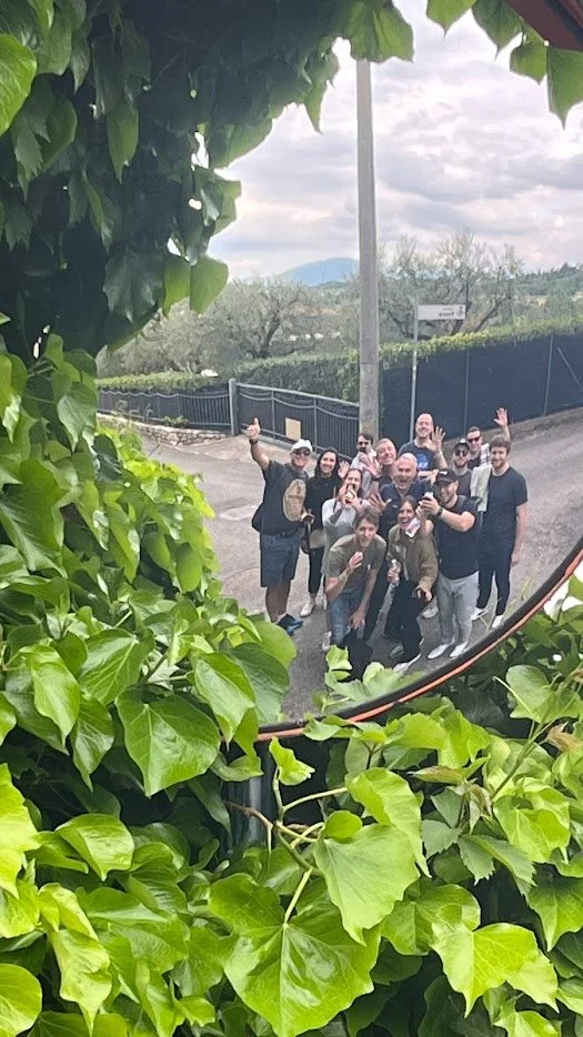 A group of people posing for a photo outdoors, seen through a mirror surrounded by green leaves, with a cloudy sky and distant mountains in the background.