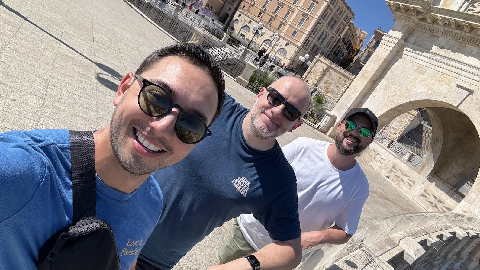 Three men taking a selfie outdoors with historic stone architecture and a cityscape in the background.