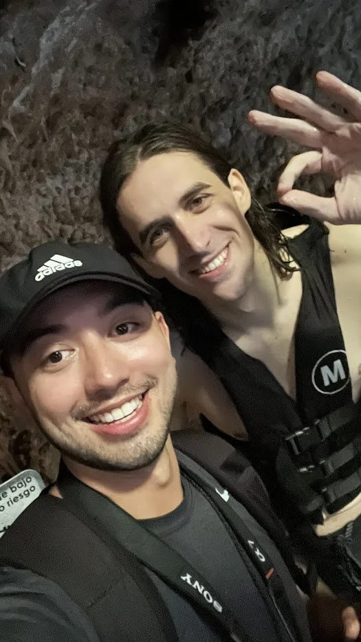 Two young men taking a selfie inside a dark rocky tunnel, smiling, with one waving at the camera.