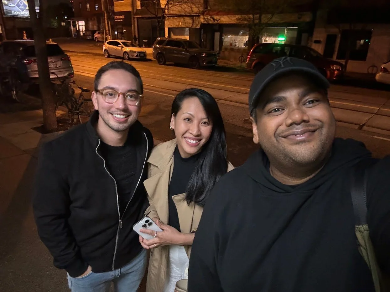 Three friends taking a selfie on a city sidewalk at night, with parked cars and tram tracks in the background.