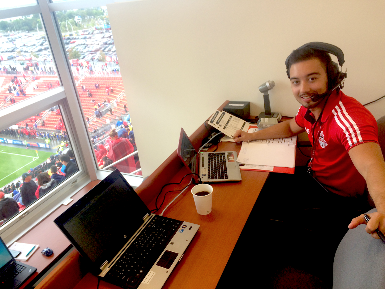 A sports commentator wearing a red Toronto FC jersey with a headphone microphone is sitting at a desk with two laptops, notebooks, a cup of coffee, and a microphone, overlooking a soccer stadium from a booth.