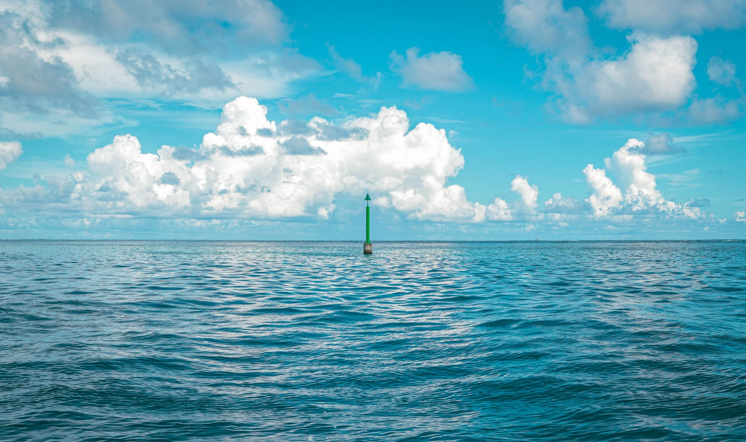 Open ocean with calm blue waters and a sky with scattered white clouds, a green navigation buoy with an arrow pointing upward in the water.