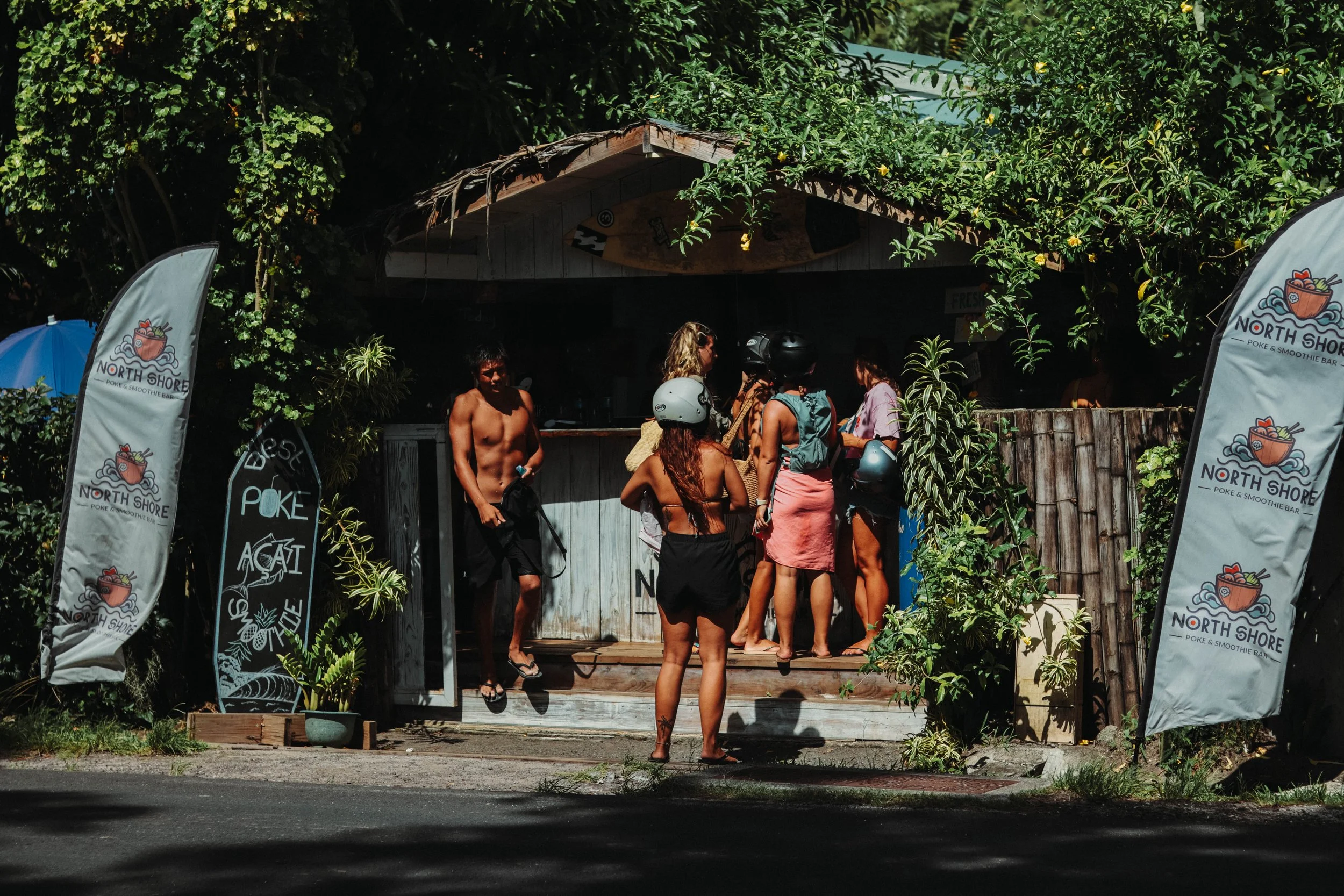 A group of people wearing helmets and swimsuits gathered outside a small beachside shack with signs that read 'North Shore' and 'Poke & Smoothie Bar,' surrounded by green foliage.