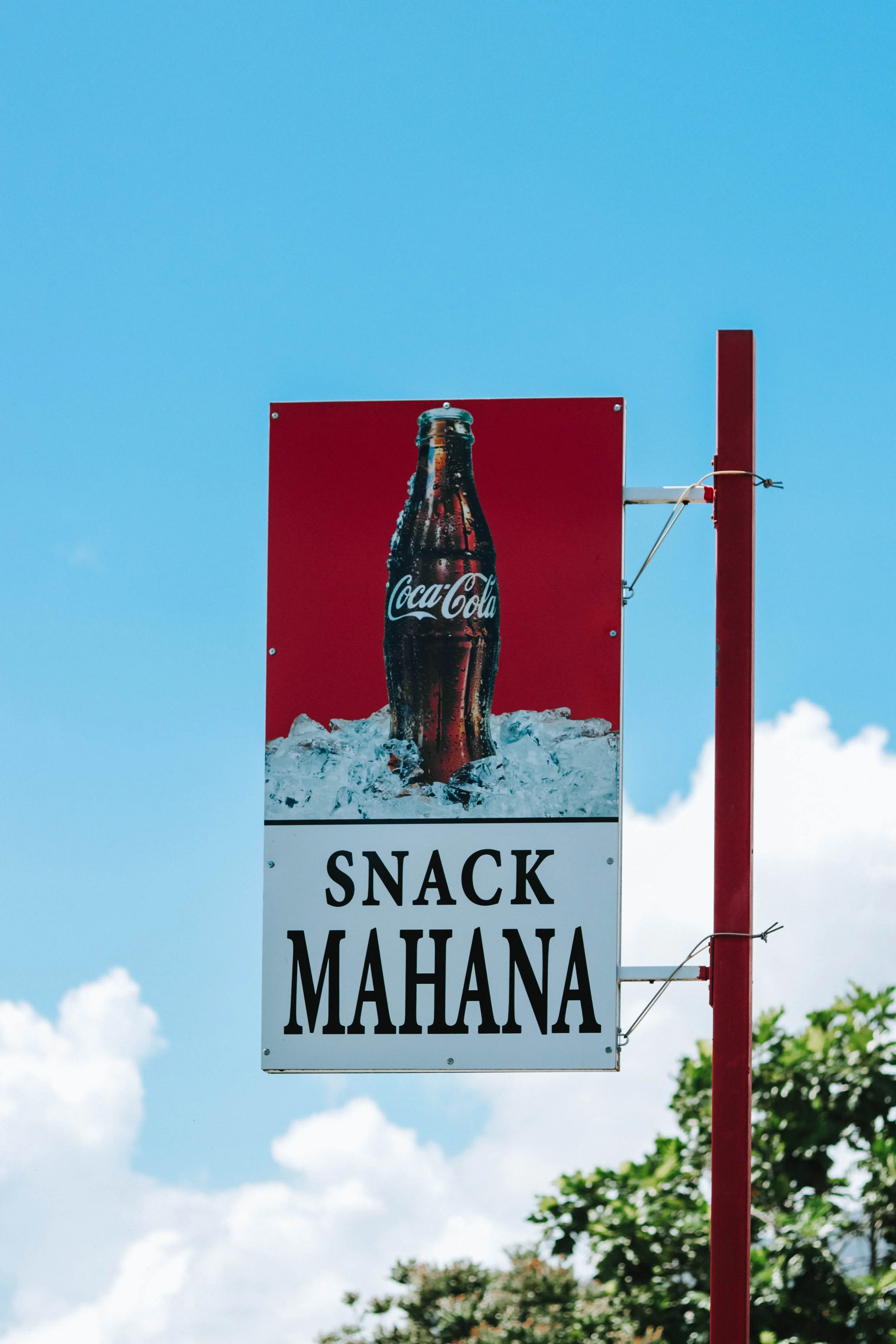 Outdoor advertising sign for Coca-Cola featuring a bottle of soda on ice, with the text 'SNACK MAHANA'.