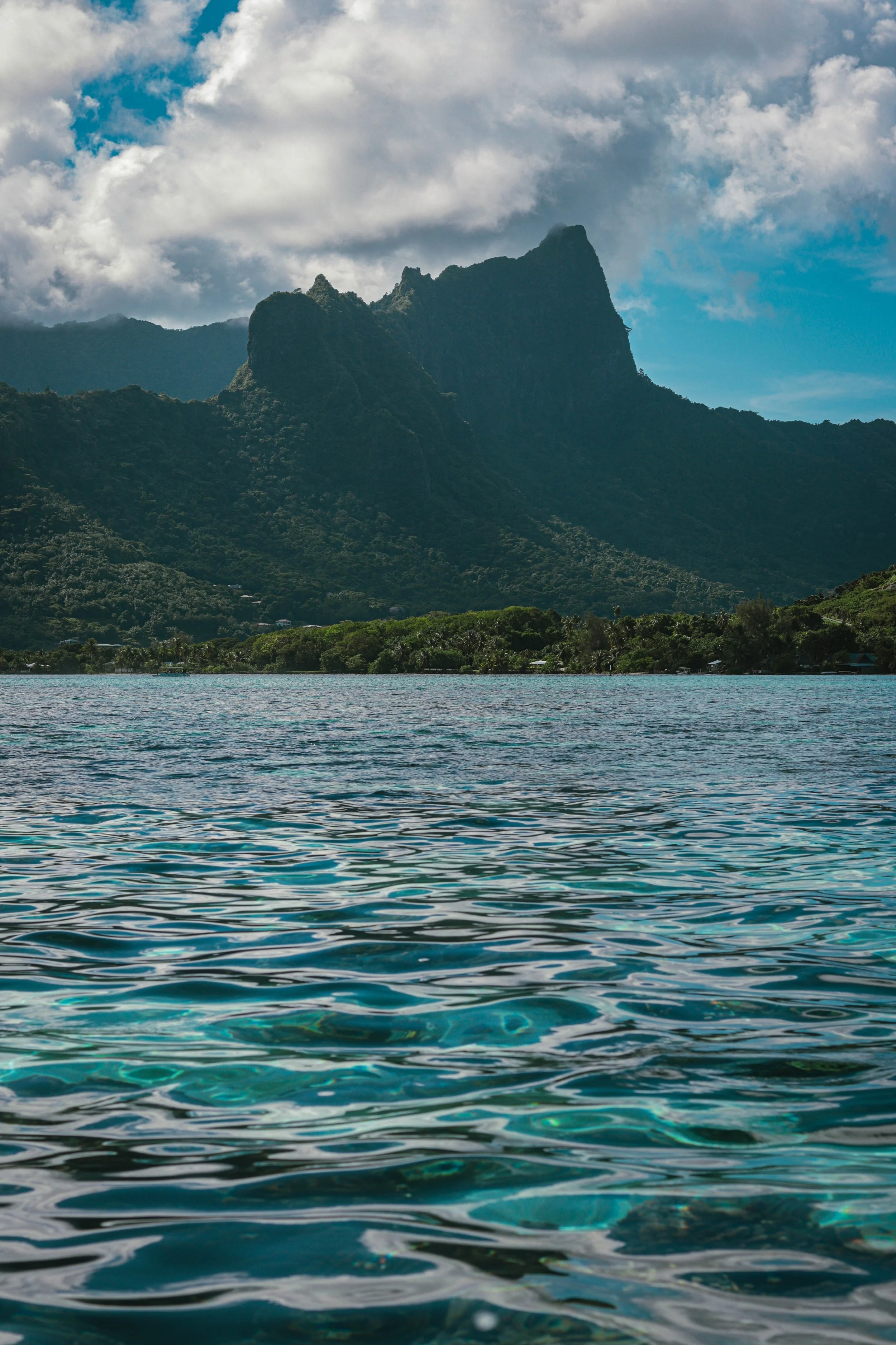 Mountains and lush greenery with a body of clear, rippling water in the foreground under a cloudy sky.