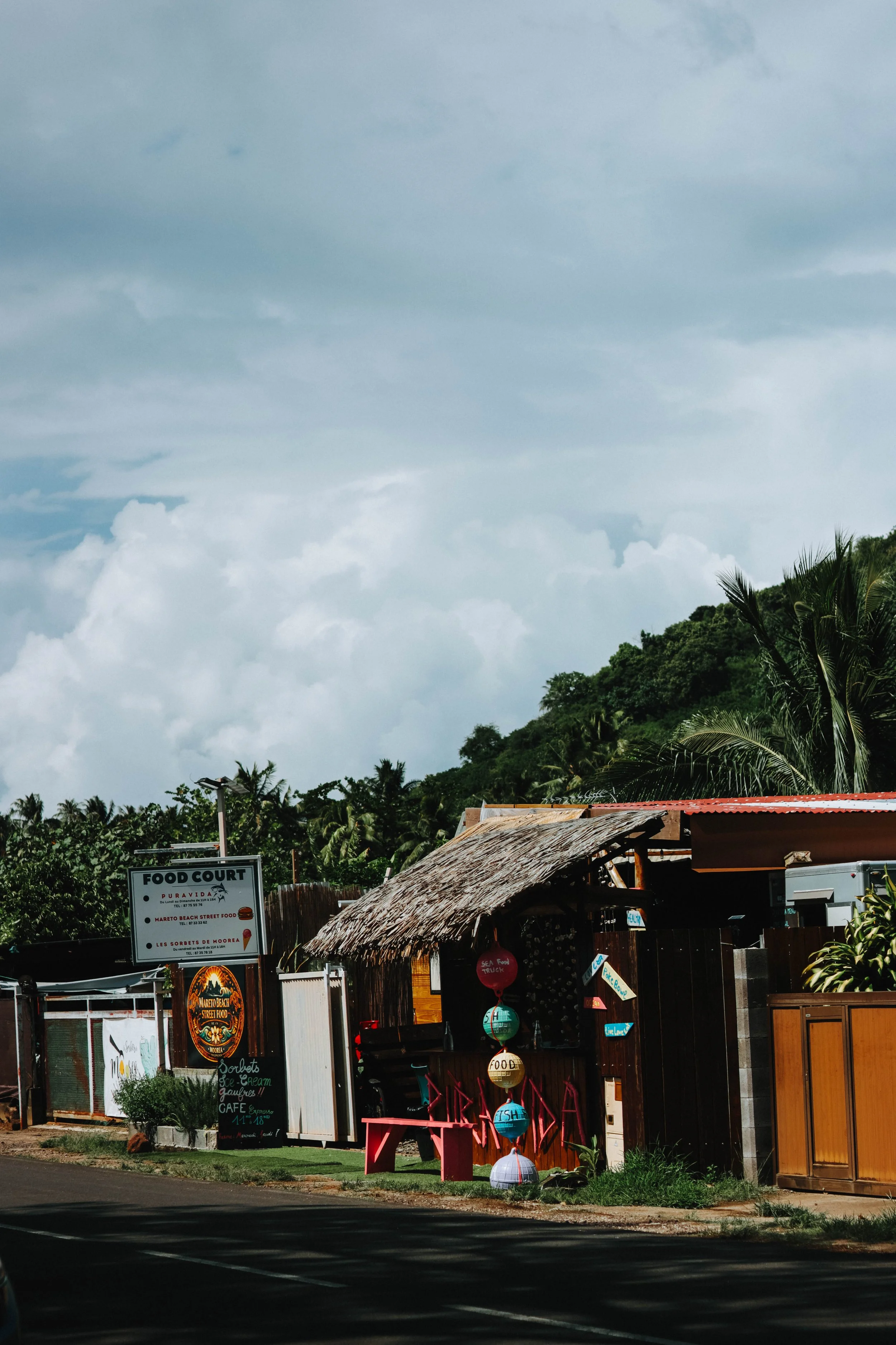 A small food stand with a thatched roof and colorful hanging lanterns in front of a tropical hillside with palm trees, under a cloudy sky.