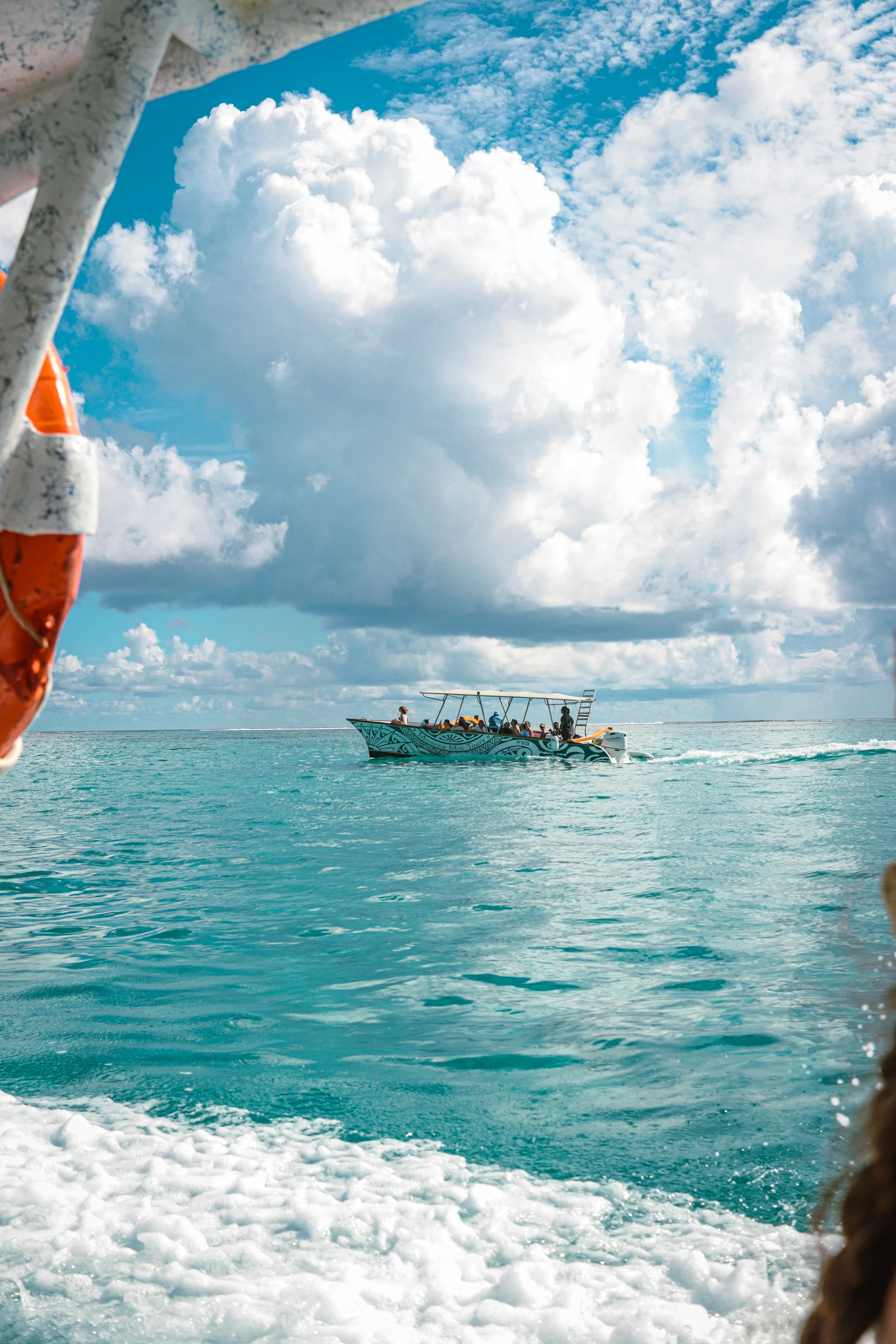 A boat with a decorated blue and white design on the side is cruising on clear turquoise water under partly cloudy skies, with a life ring visible in the foreground.