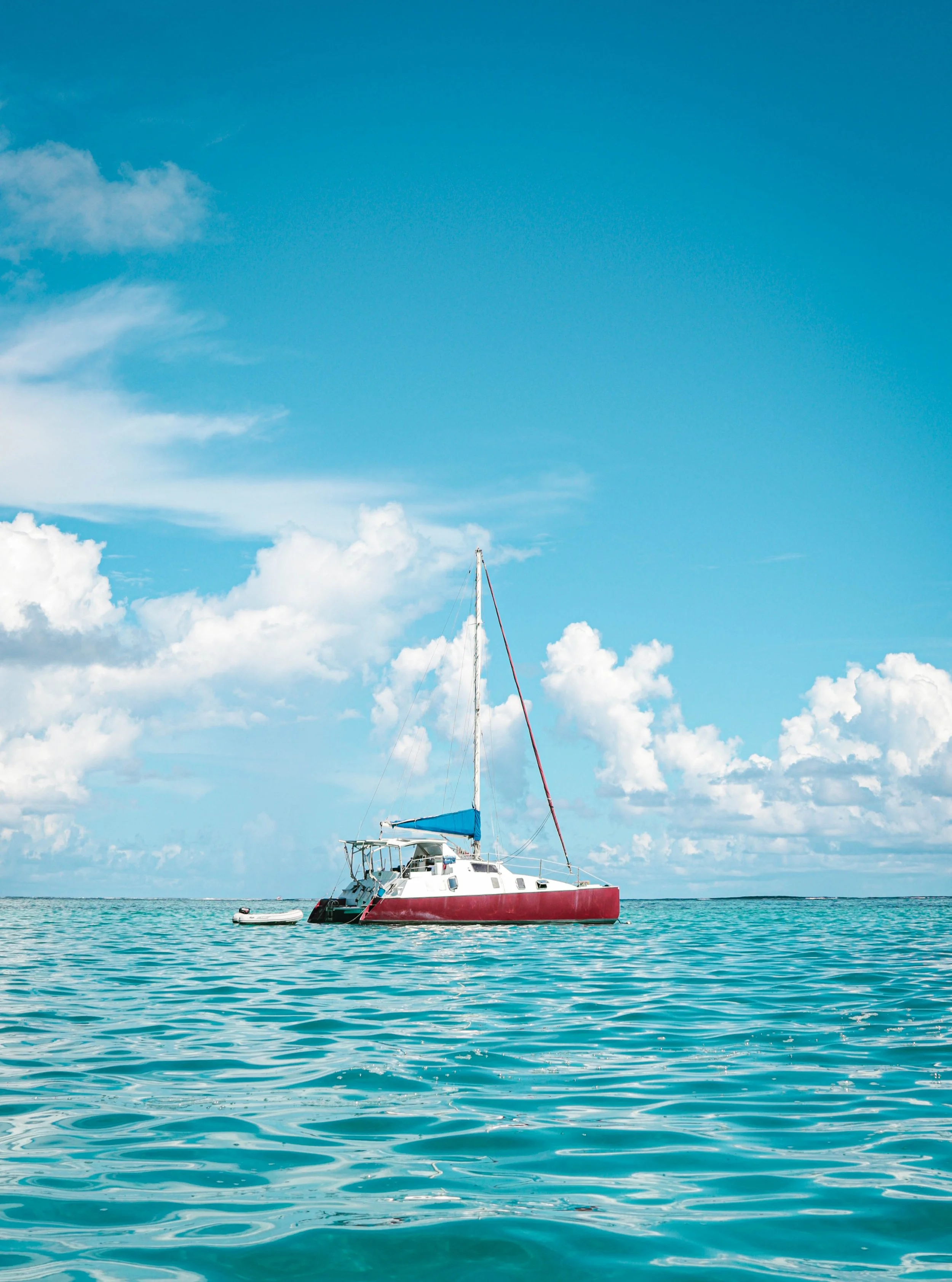 A sailboat with a red hull, blue sail cover, and a small inflatable dinghy attached to it, floating on calm turquoise water under a partly cloudy blue sky.