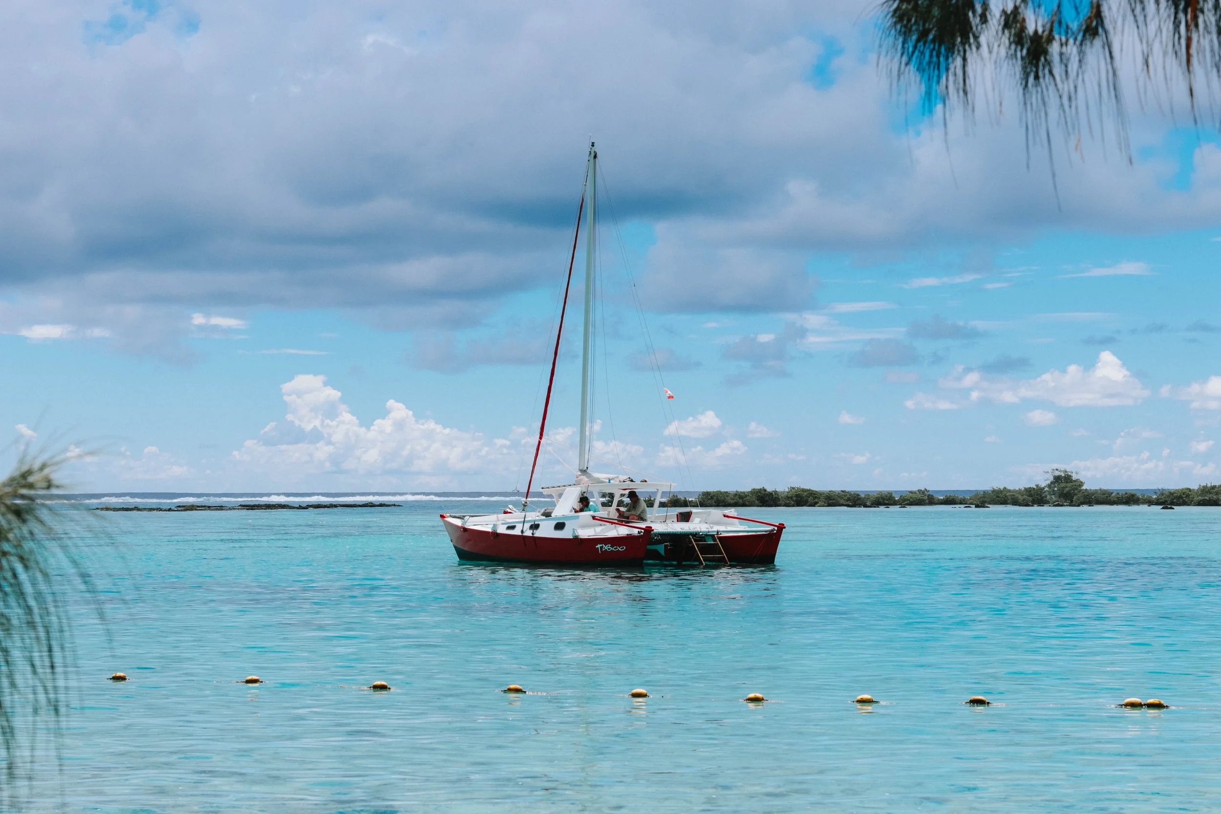 A red and white sailboat floating on turquoise water under a partly cloudy sky, with distant land and trees on the horizon.