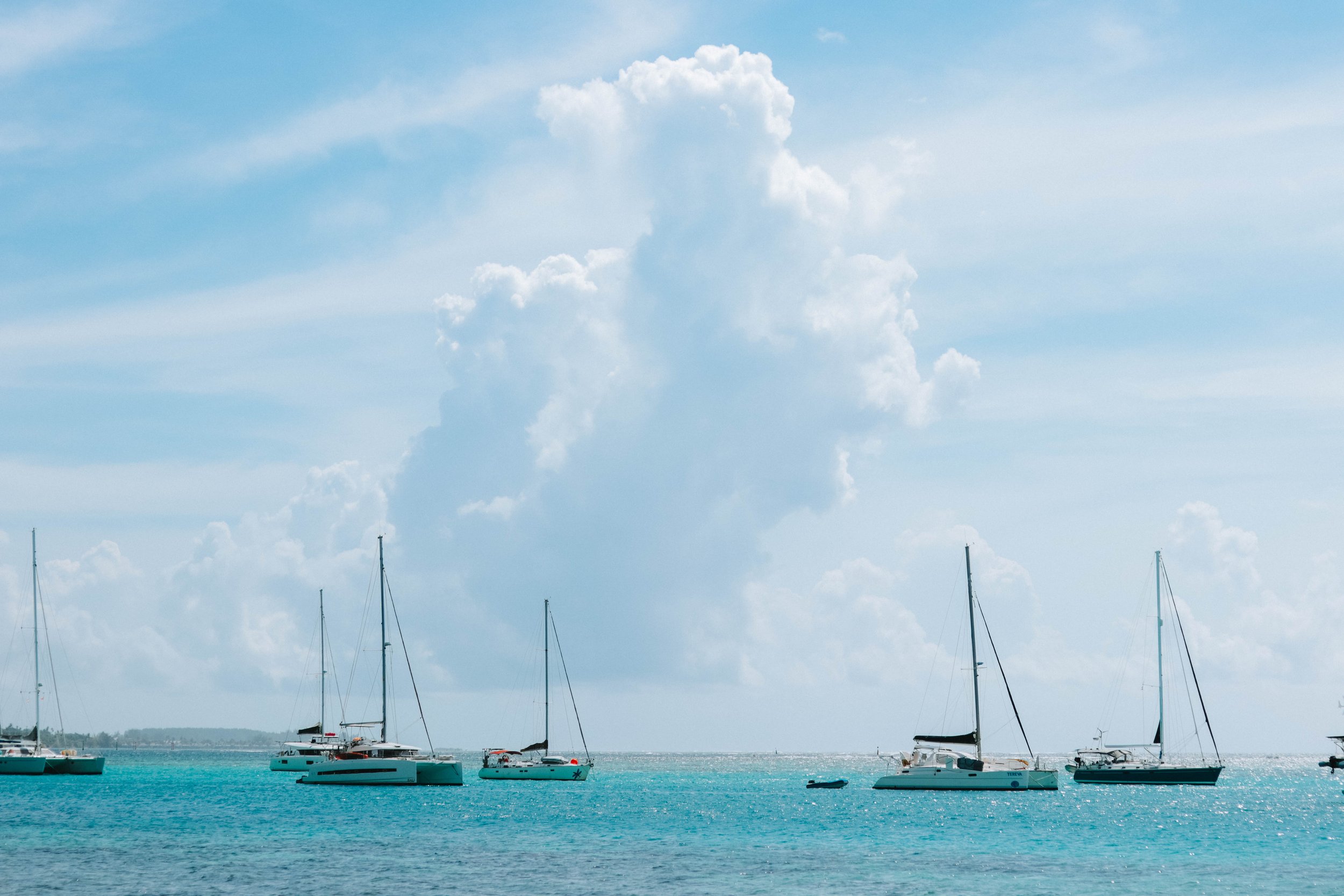 Several sailboats anchored on calm turquoise water with a distant shoreline and a partly cloudy sky.
