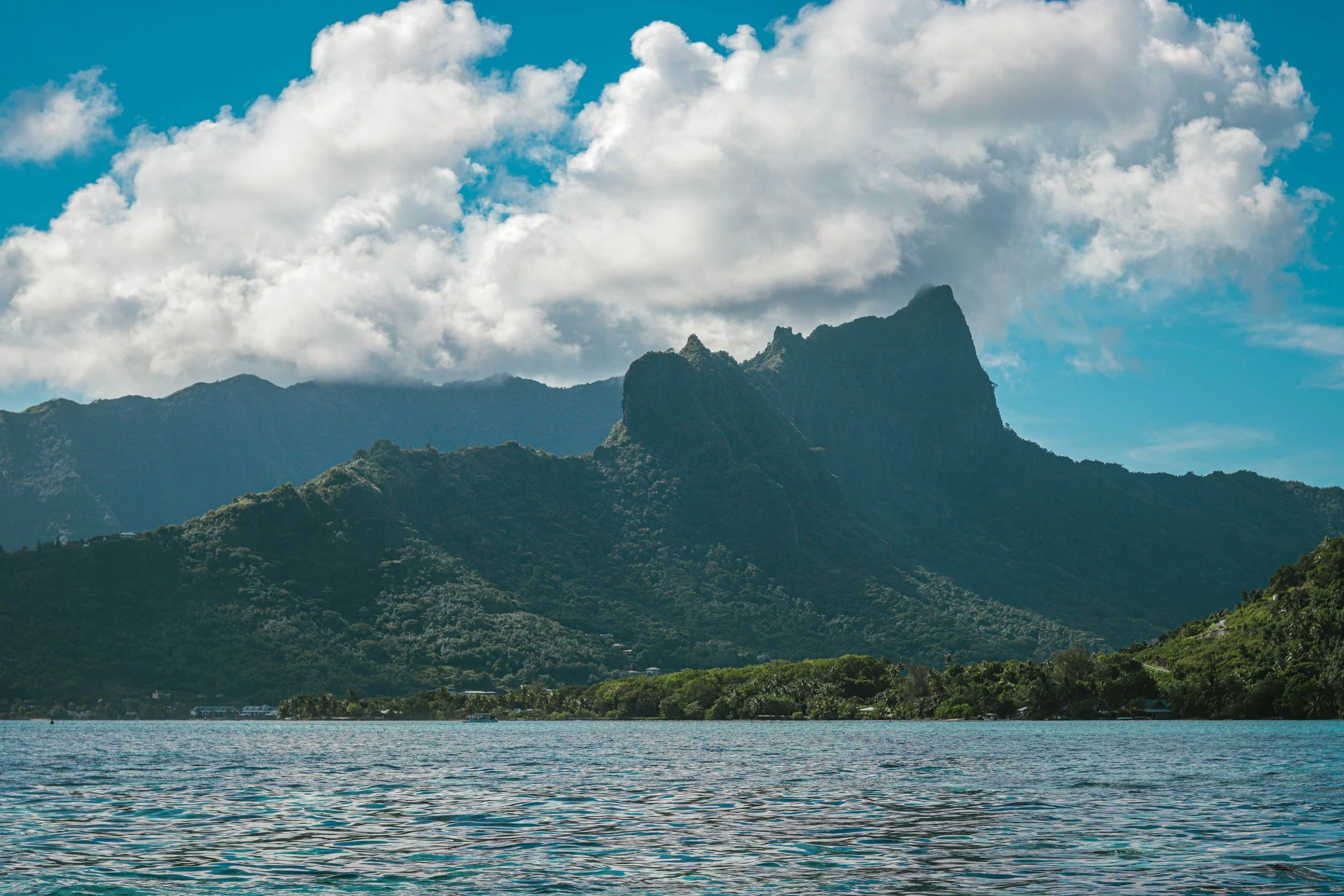 A scenic view of a mountain range with lush green slopes, a body of water in the foreground, and a partly cloudy sky with large white clouds.