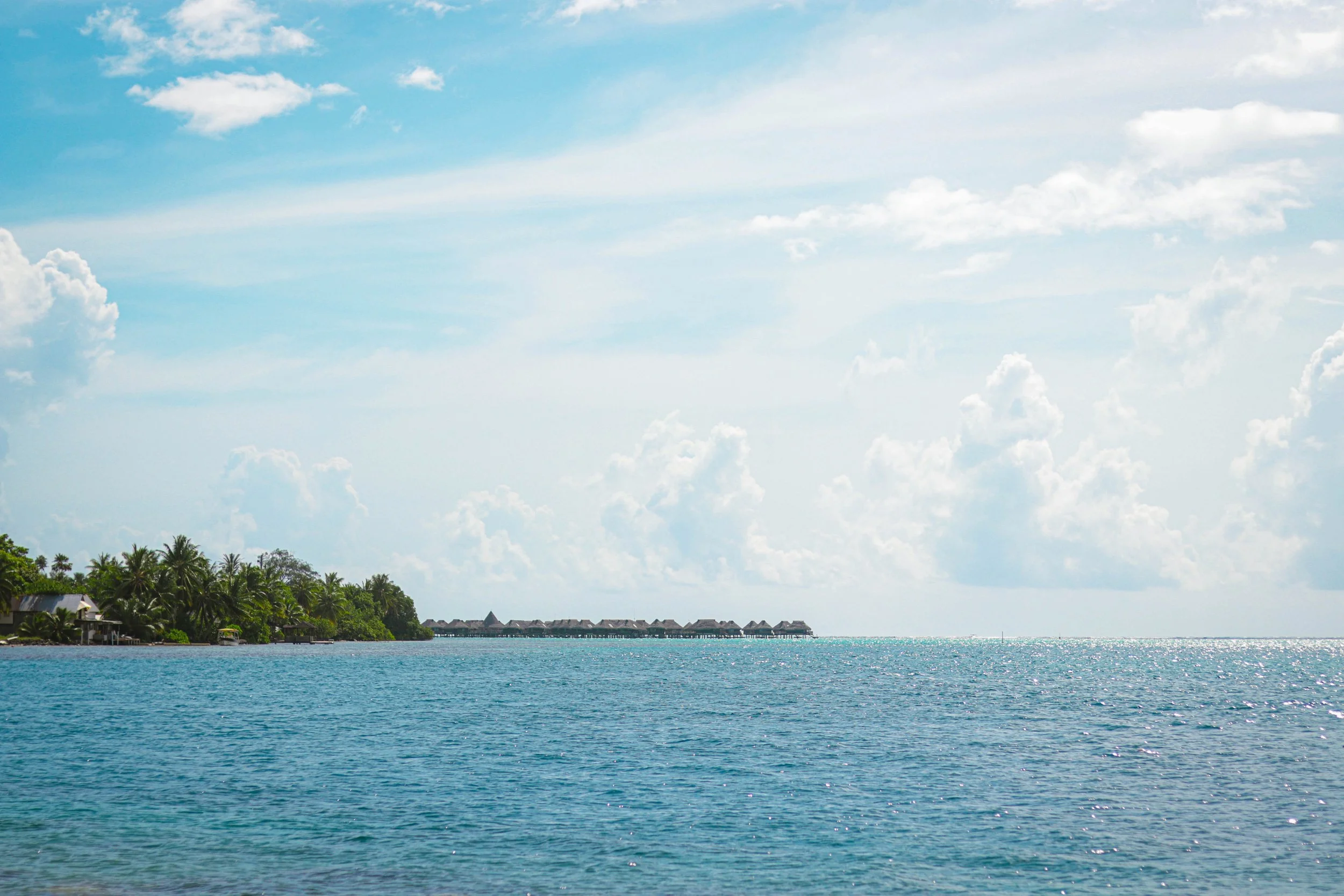 Tropical ocean with overwater bungalows in the distance, lush greenery on the shoreline, and a bright blue sky with some clouds.
