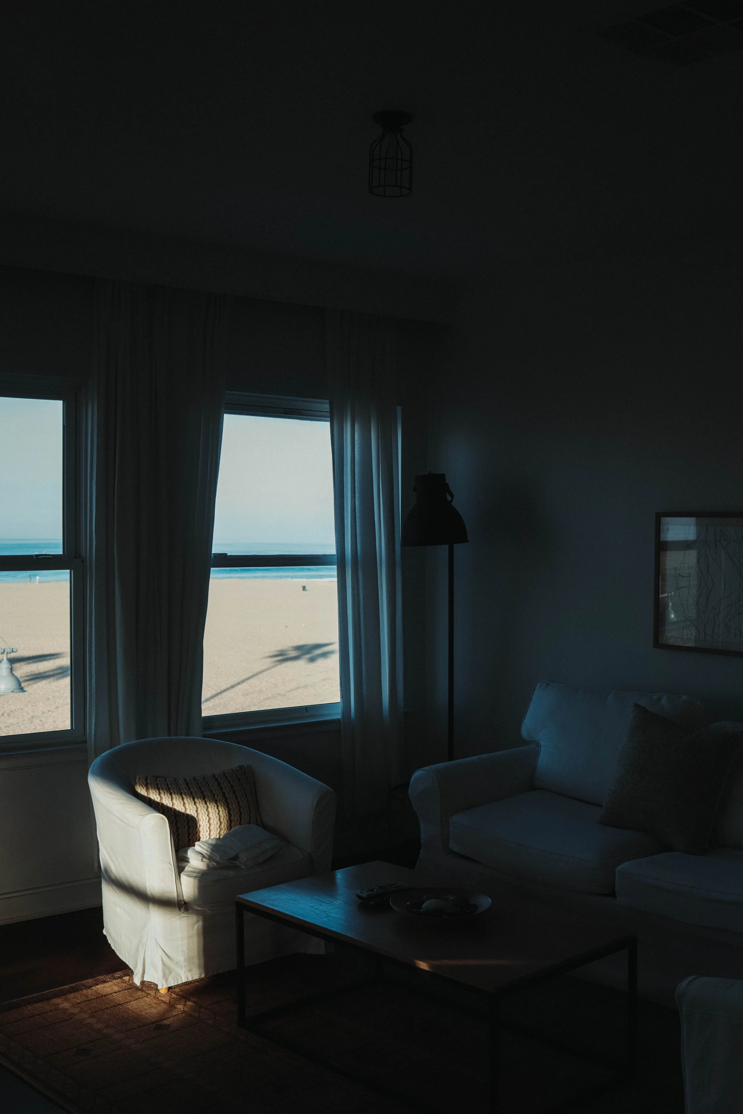 A darkened living room with sunlight coming through windows, showing a view of the beach and ocean outside.