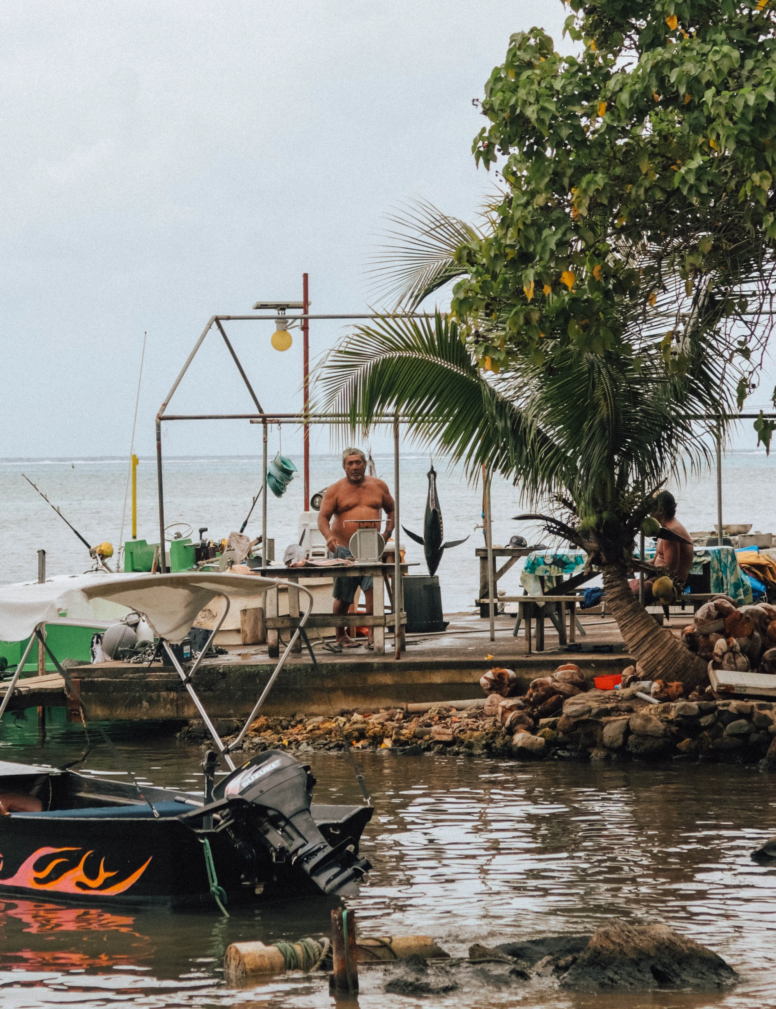 A man is standing shirtless on a pier by the water, surrounded by fishing equipment and boats, with a tree providing shade nearby.