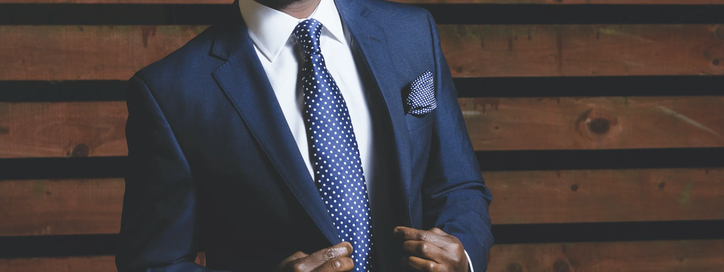 Man in a navy suit, white shirt, and polka dot tie with matching pocket square, standing against a wooden background.
