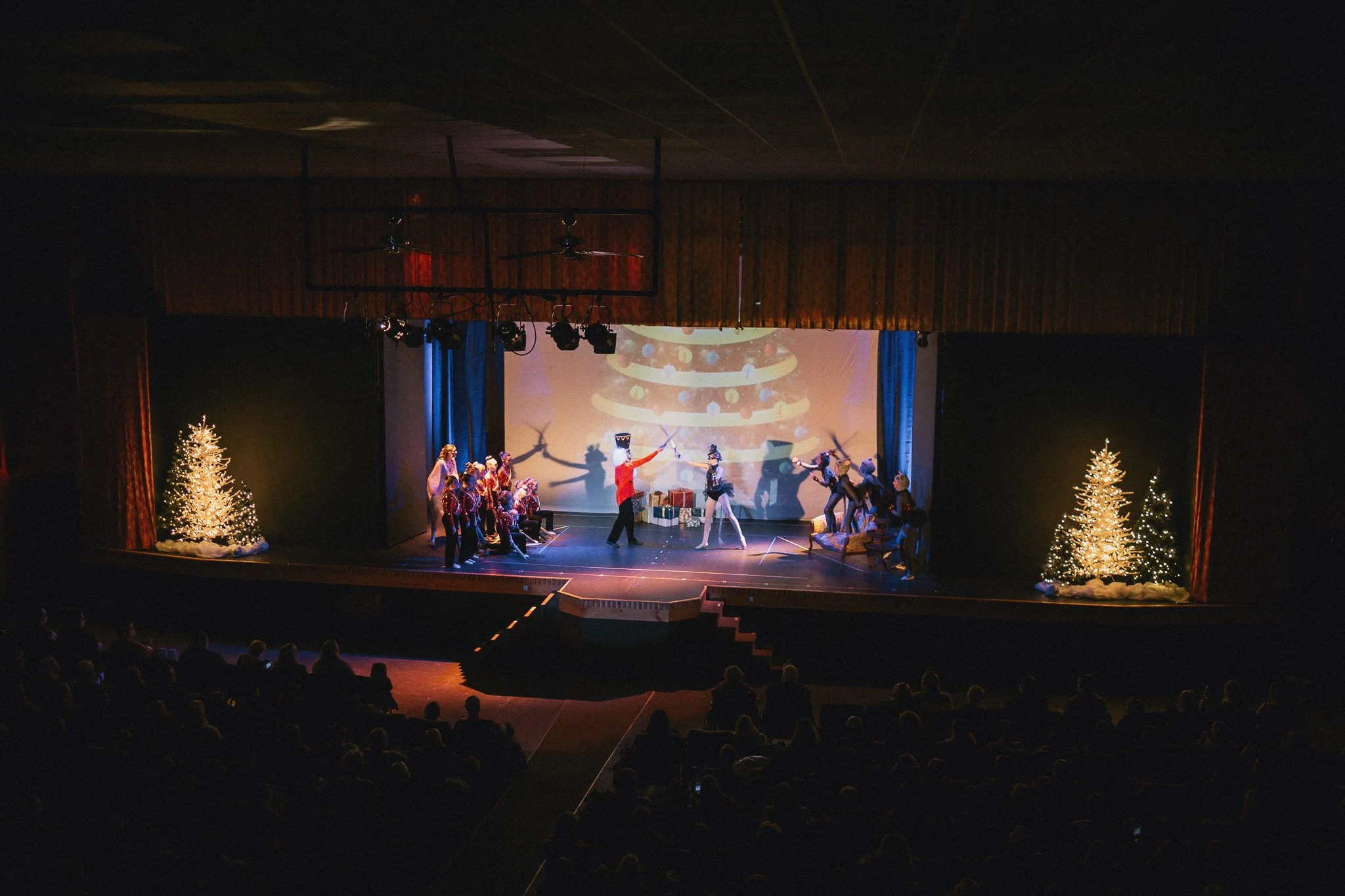 A stage production of a Christmas-themed performance, featuring actors dressed in costumes, Christmas trees with lights, and a backdrop with a decorated Christmas tree projection.