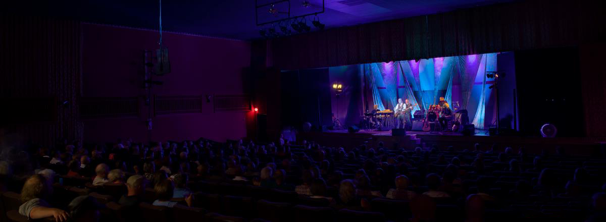 Concert stage with musicians performing under colorful lighting, audience seated in a darkened theater watching the show.