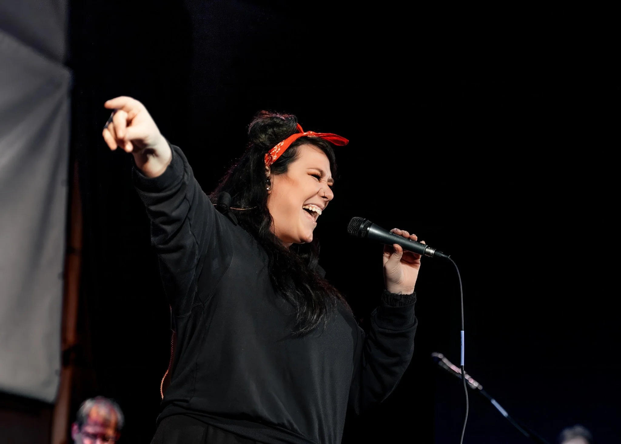 A woman with dark hair wearing a red bandana is singing passionately into a microphone on stage, with her eyes closed and face joyful.