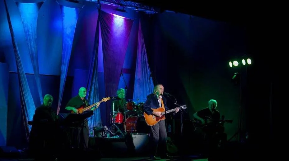 A musical band performs on stage with five members, including a lead singer playing guitar, under colorful stage lighting.