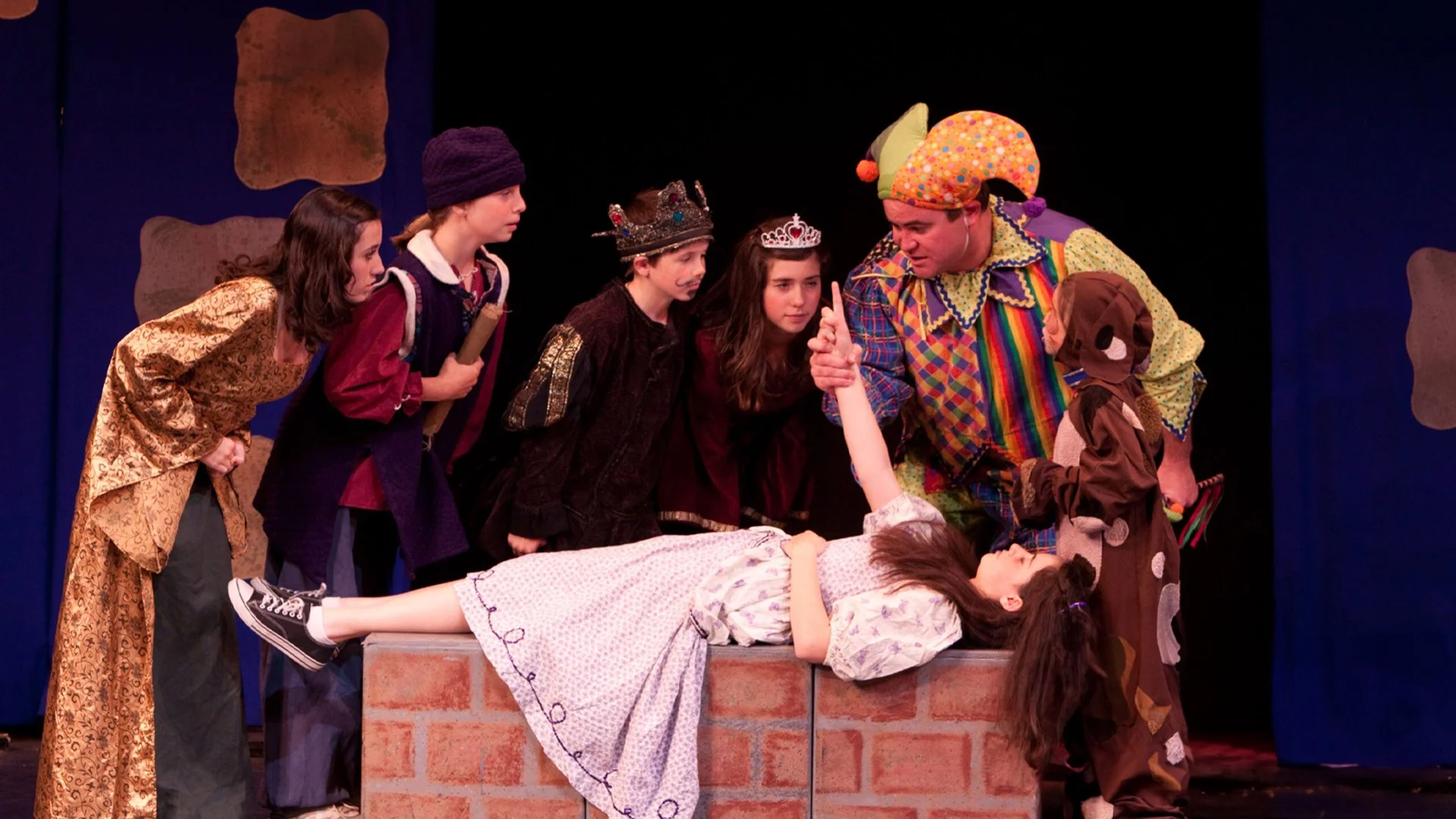 Children and adults in costumes performing a scene with a girl lying on a table as if she is asleep, while others look on and a clown character gestures.