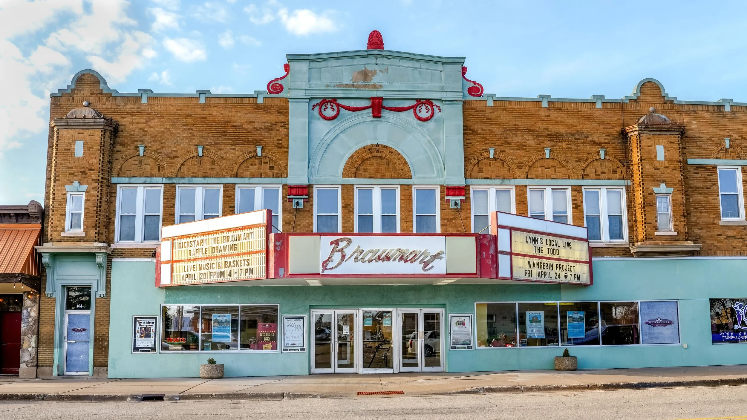 The facade of a vintage theater with a marquee showing event details, above a turquoise storefront, under a blue sky with clouds.