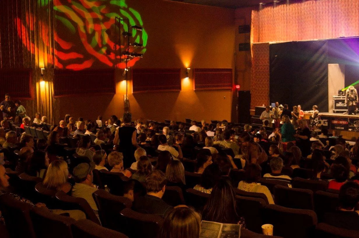 Audience sitting in theater seats watching a performance on stage with colorful lighting effects.