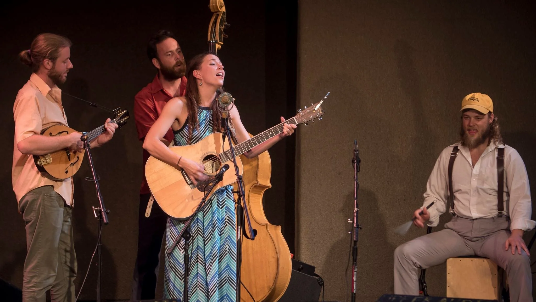 Four musicians performing on stage; three men playing instruments and a woman singing with a guitar.
