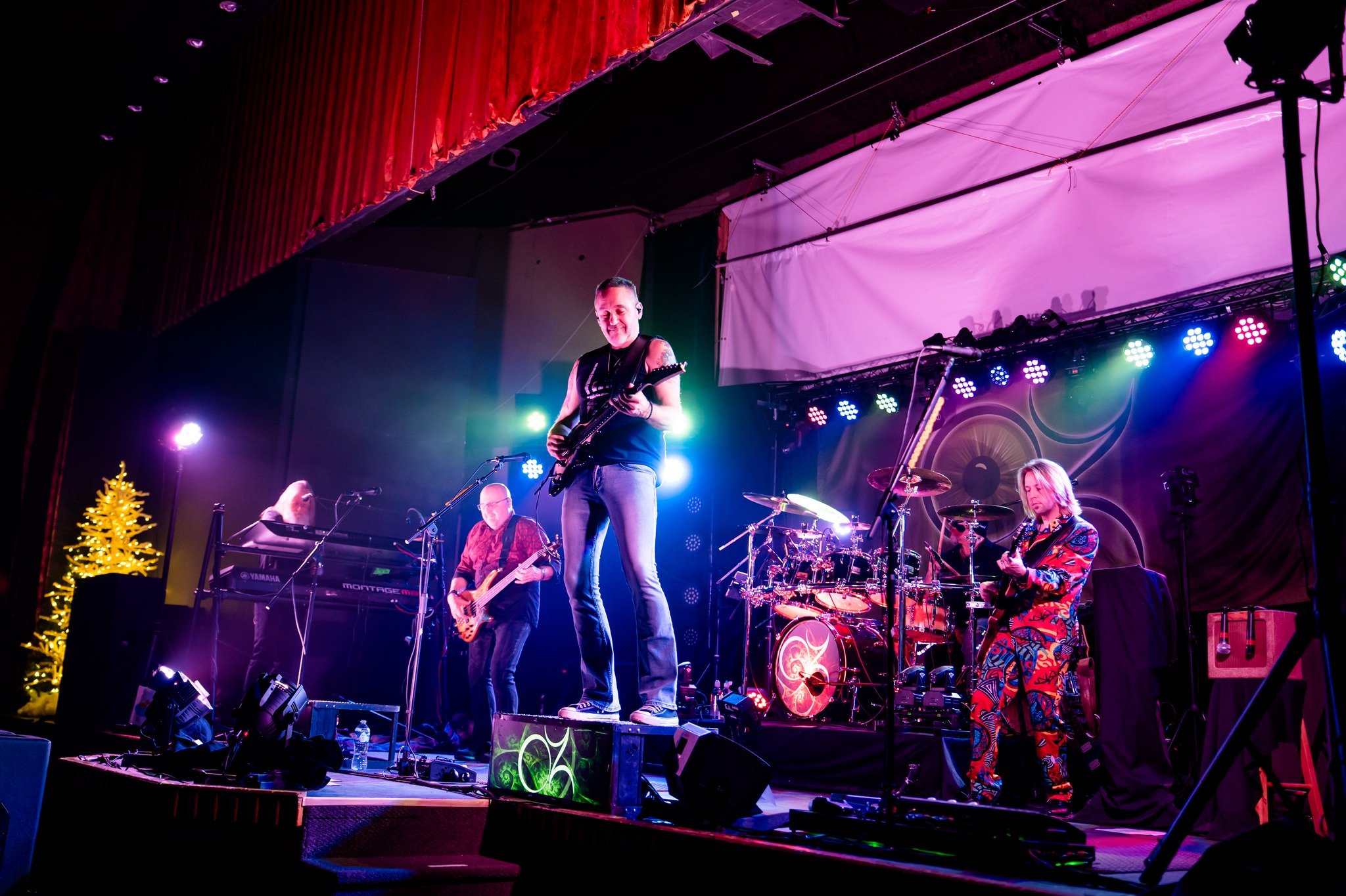 A band performing on stage with colorful lighting, including a guitarist, keyboardist, bassist, drummer, and another guitarist, with a Christmas tree on the left side of the stage.