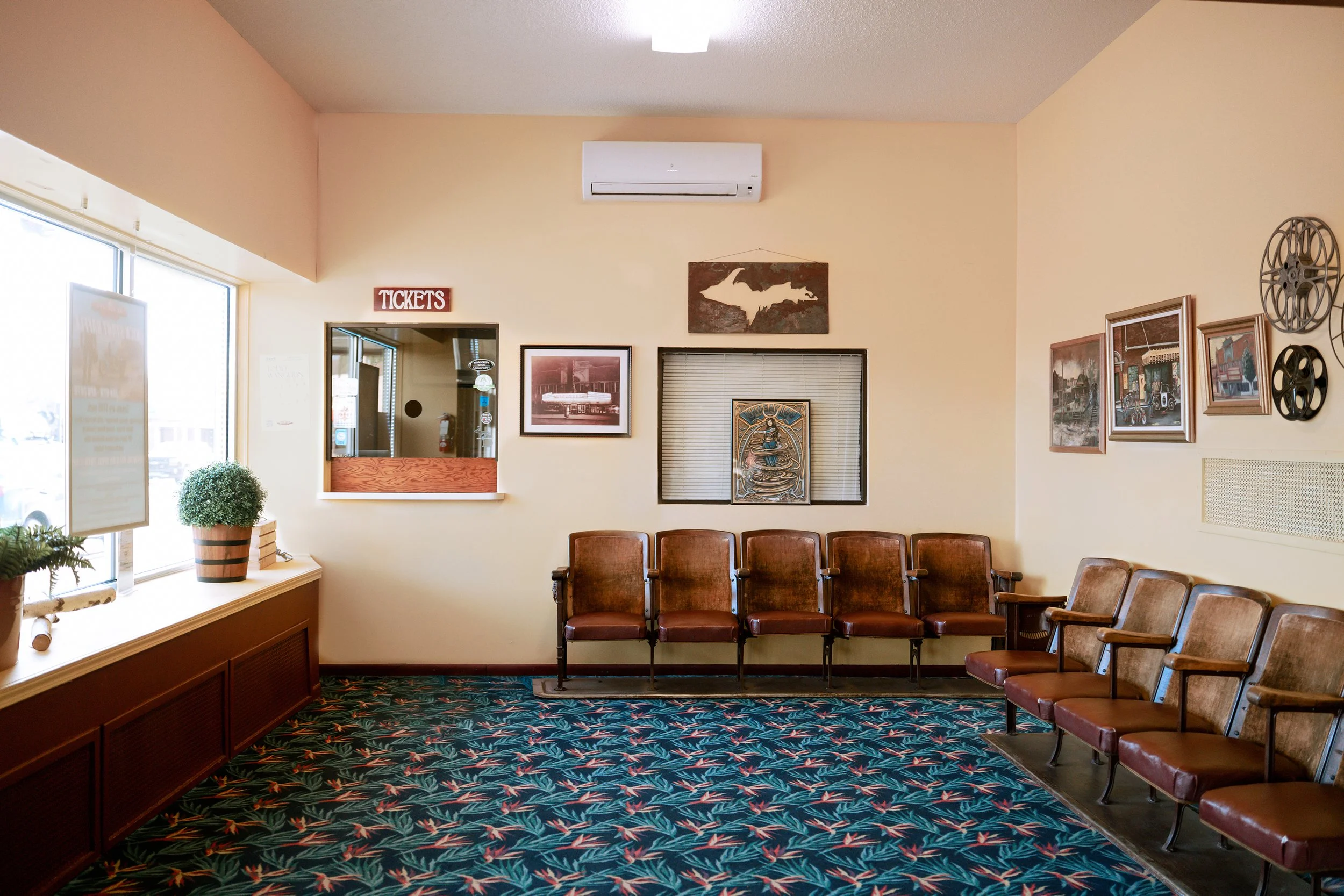 A waiting room with brown chairs arranged in a row along a wall. The room has beige walls with framed pictures and decorative items. There is a window with a small potted plant on the windowsill and a patterned carpet on the floor.