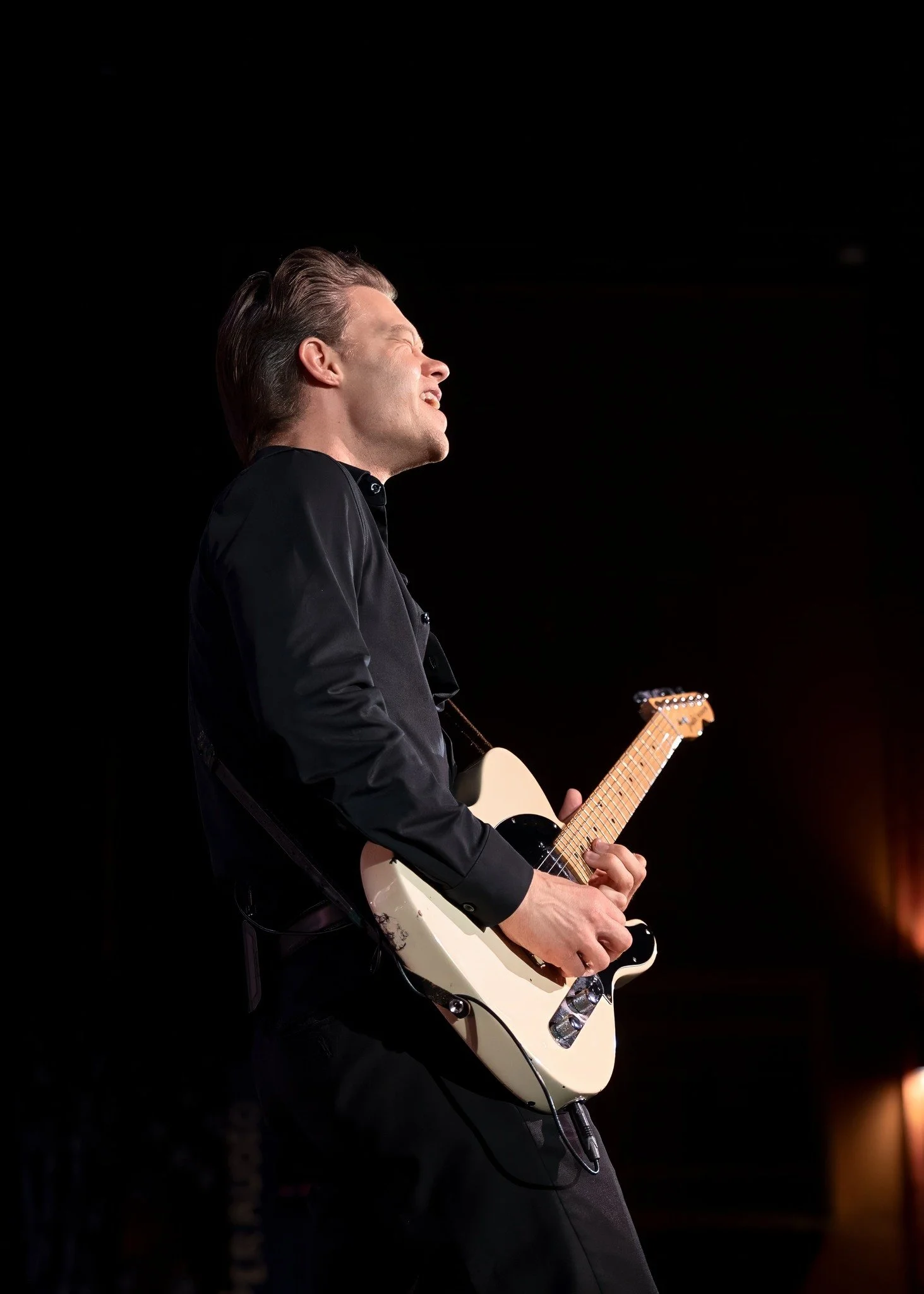 A man playing the guitar on stage, singing with his eyes closed, wearing a black shirt, against a black background.