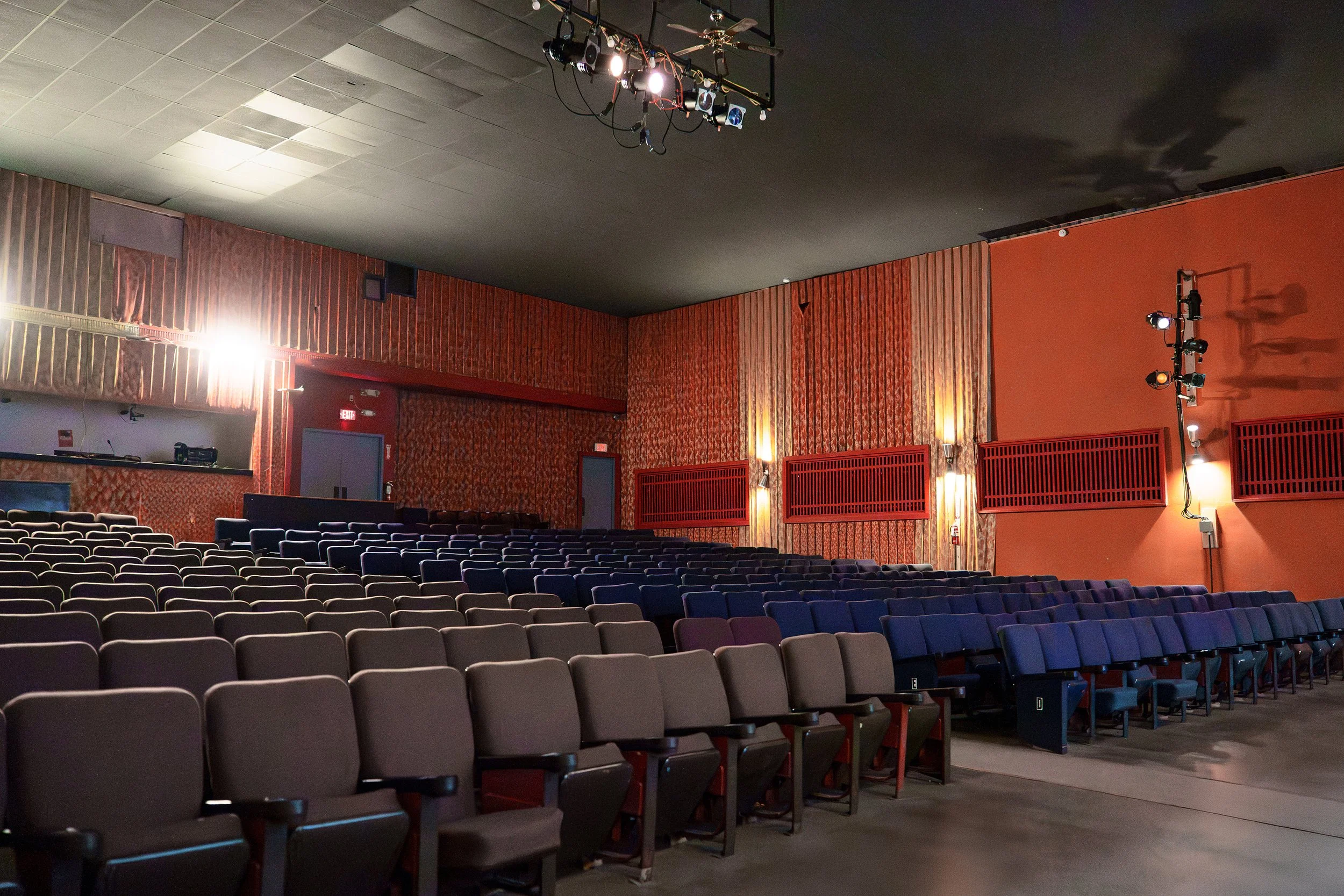 Inside of an empty auditorium with rows of gray chairs, wall-mounted lights, and a large window. Stage lighting equipment is hanging from the ceiling.