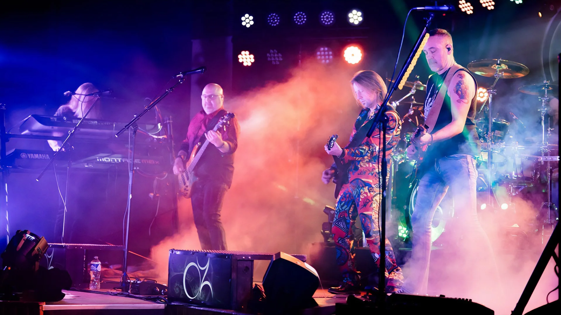OZ: TheBand performing on stage with colorful lights and fog, including keyboard, bass guitar, electric guitars, and drums.