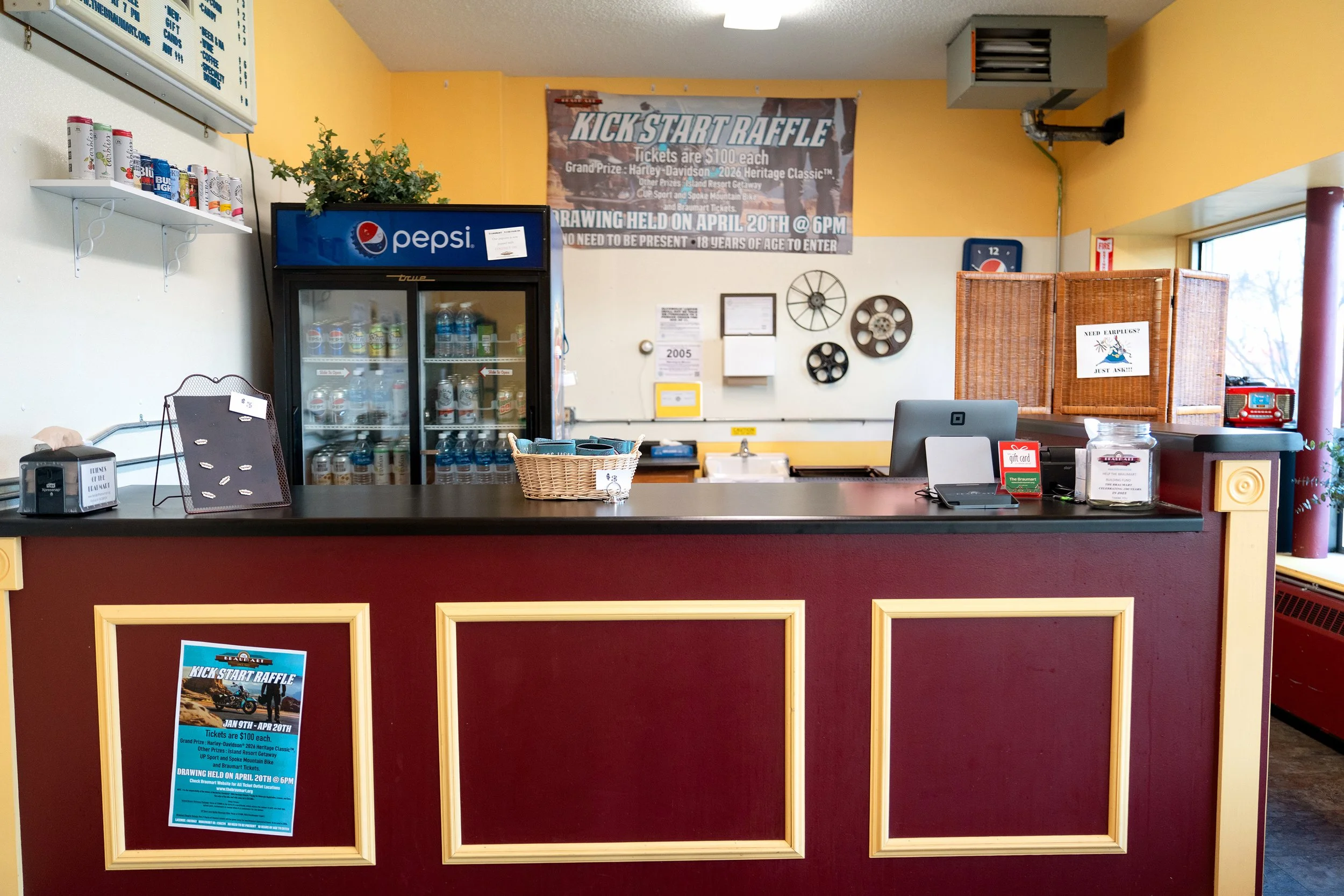 A counter area with a Pepsi fridge behind it, a basket, a computer, a sign about earplugs, and posters and decorations including a large banner about a kick start raffle and other wall decor.