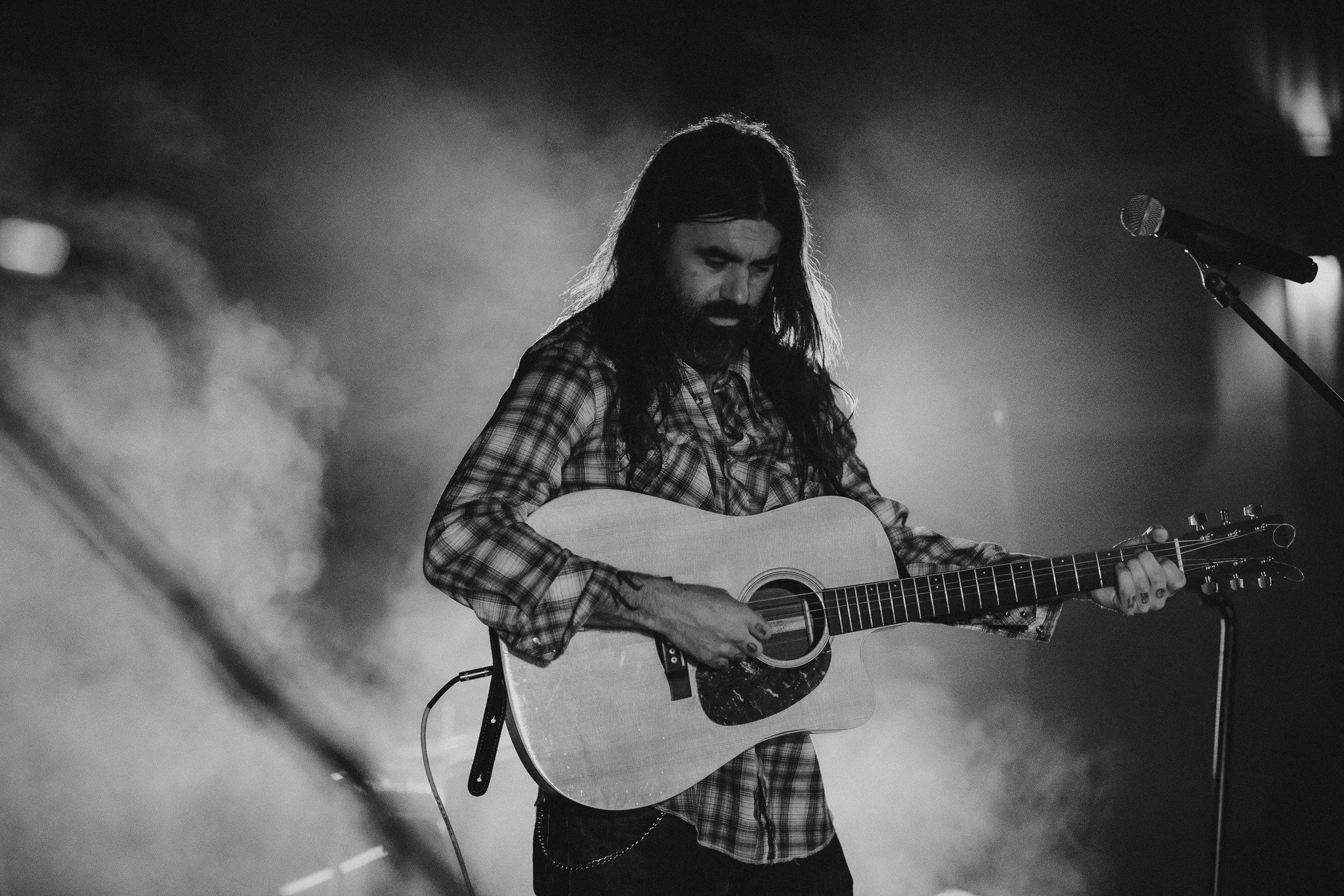 A man with long hair and a beard plays an acoustic guitar on stage, wearing a plaid shirt with a microphone nearby.