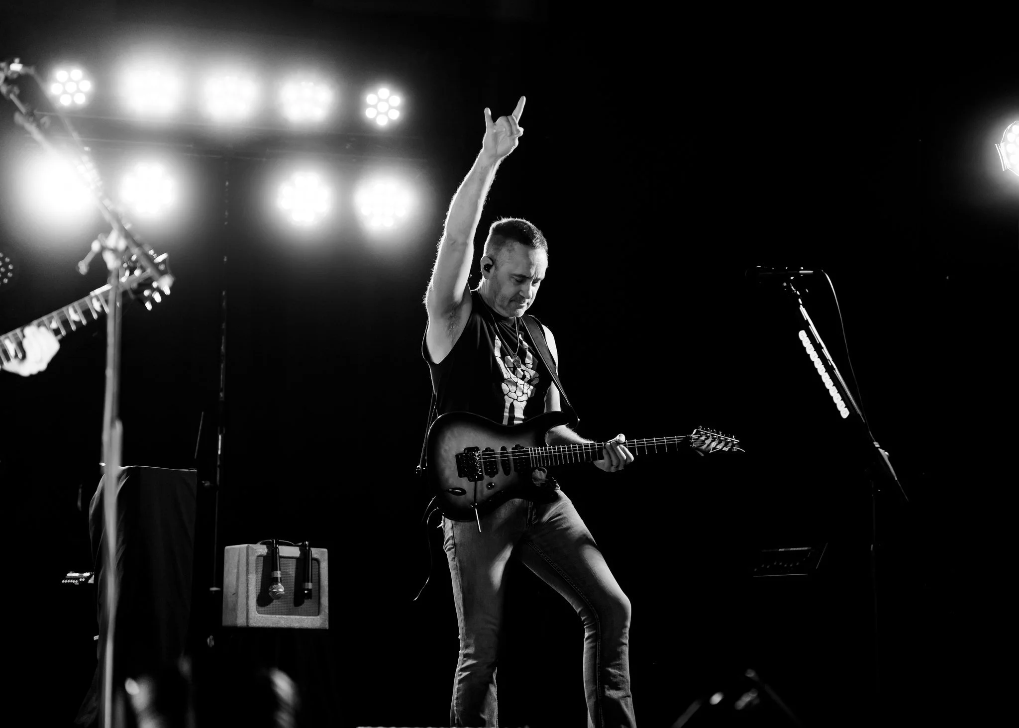 Musician playing an electric guitar on stage with stage lights overhead, black background, and a small amplifier nearby.