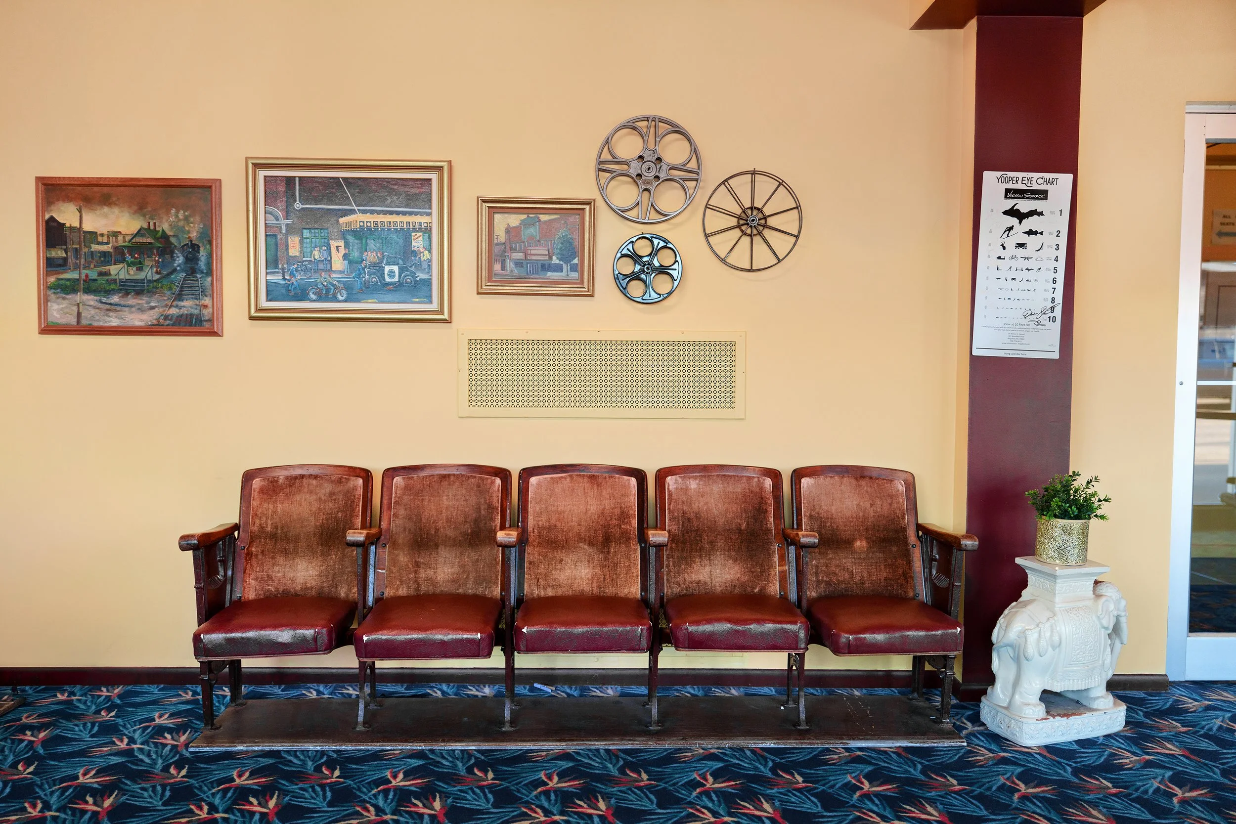 A row of five reddish-brown vintage chairs with wooden armrests, set against a yellow wall with artwork and metal wall decor. To the right, there is a white side table with a decorative elephant sculpture and a potted green plant. A large sign with an eye chart and cartoon images is mounted on a dark red pillar near a doorway.