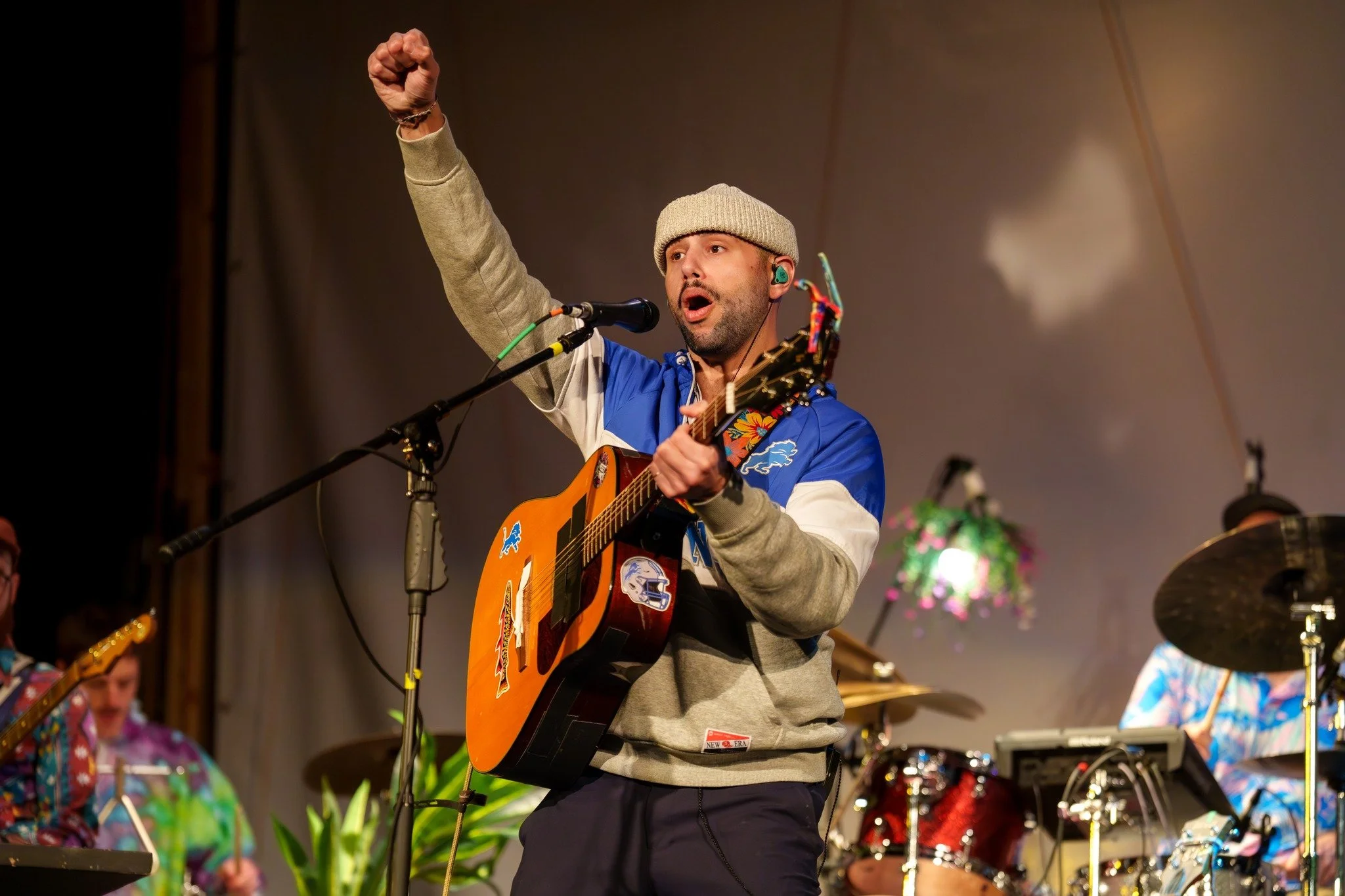 A man playing guitar and singing into a microphone during a live performance, raising his fist in the air. Other musicians are visible in the background.