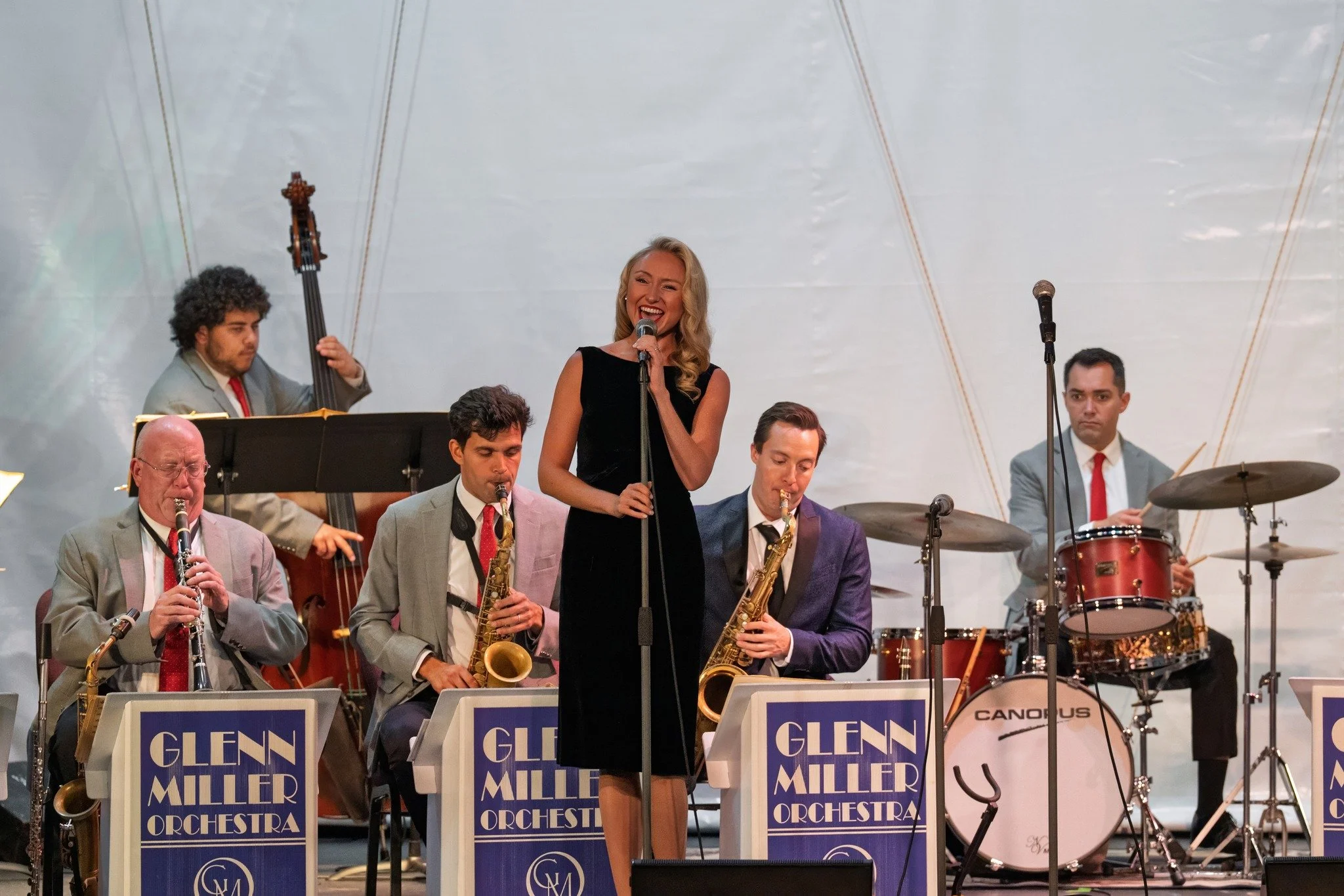 A musical performance featuring a woman singing into a microphone, surrounded by musicians playing instruments such as saxophones, drums, and a double bass, with music stands labeled 'Glen Miller Orchestra'.