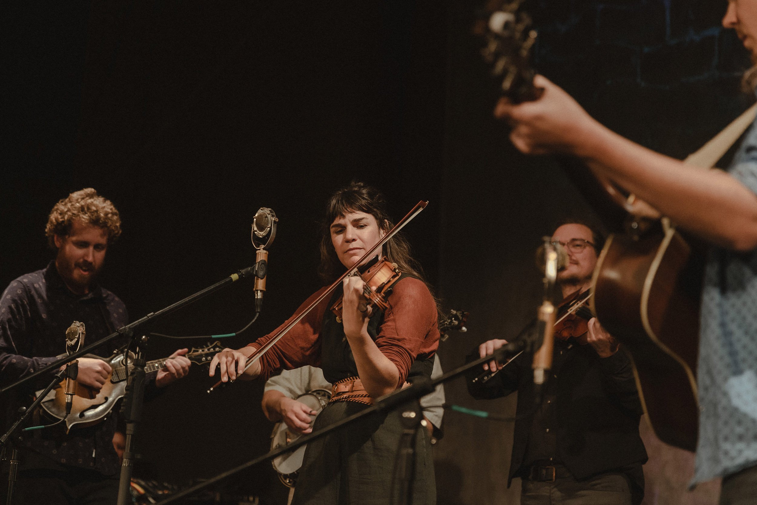 Group of musicians playing instruments, including a woman with a violin, in a dimly lit setting.