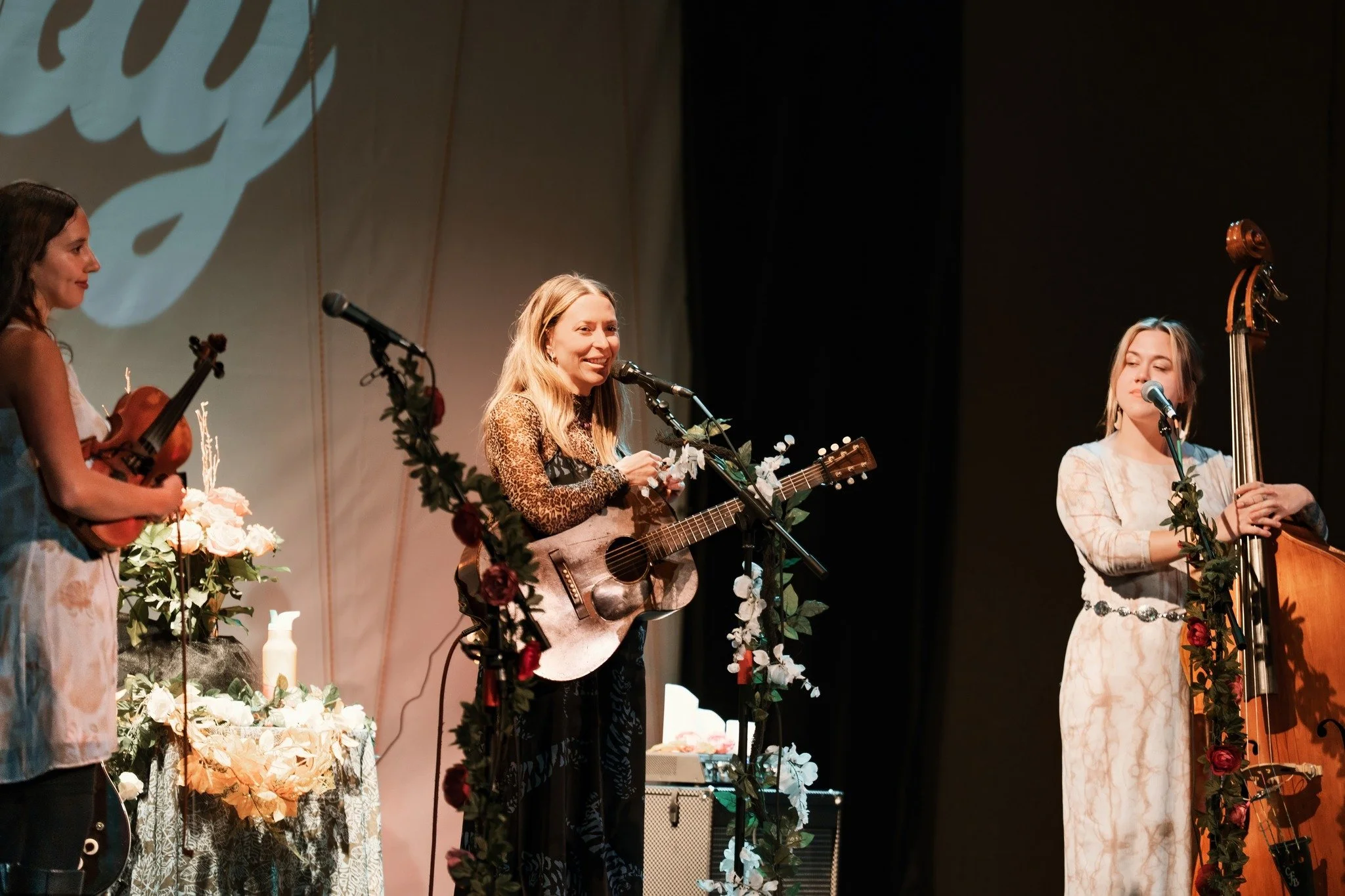 Three women perform on stage with musical instruments, surrounded by floral decorations and a backdrop with a logo, under stage lighting.