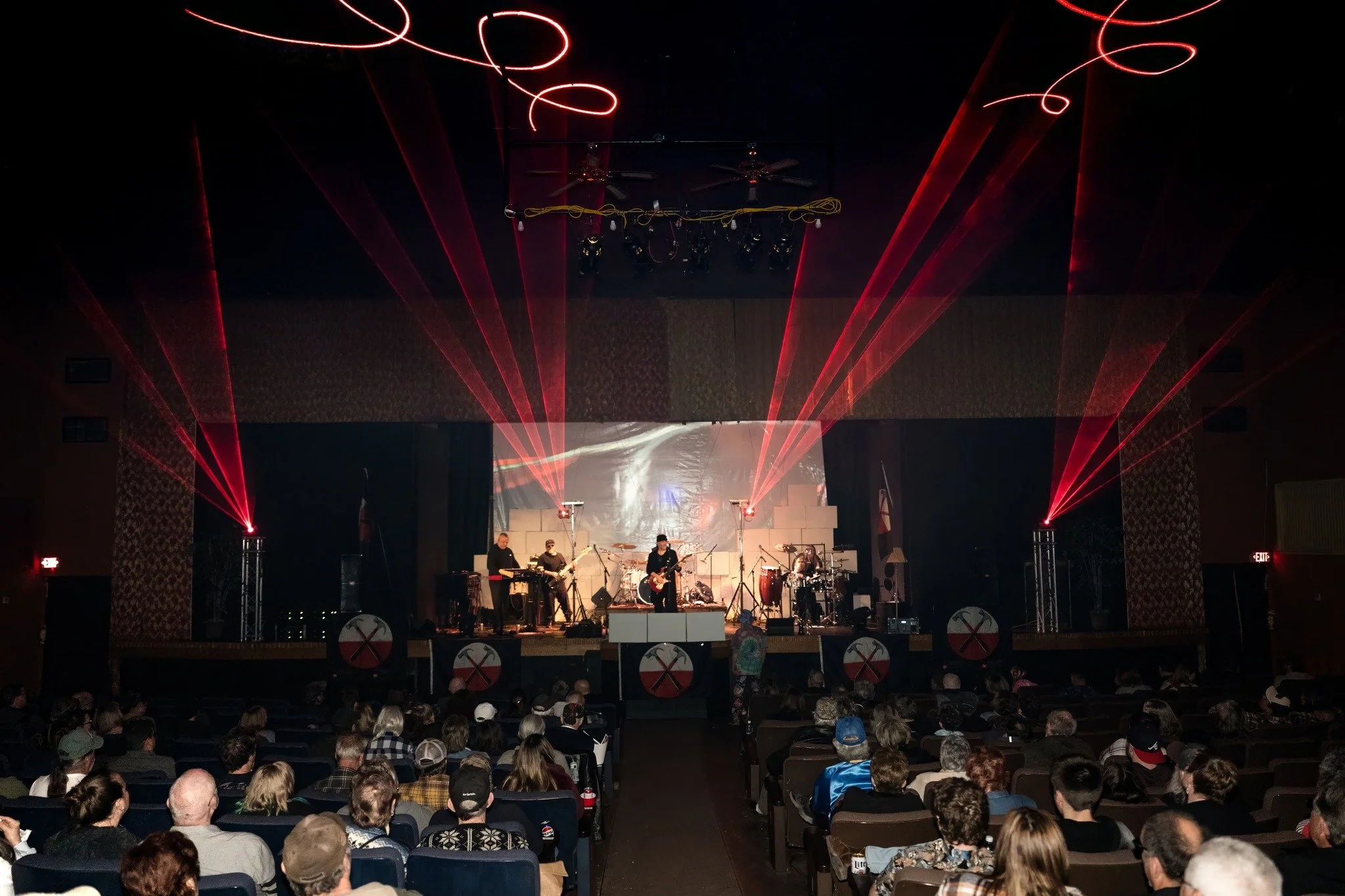 A live music concert with a band performing on stage, illuminated by red laser lights. Audience seated in front of the stage watching the performance.
