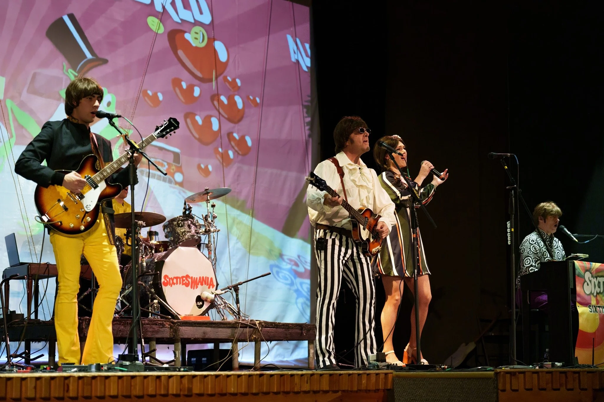 A group of musicians performing on stage with guitars and drums, featuring a colorful background with the word 'SIXTIESMANIA' visible.