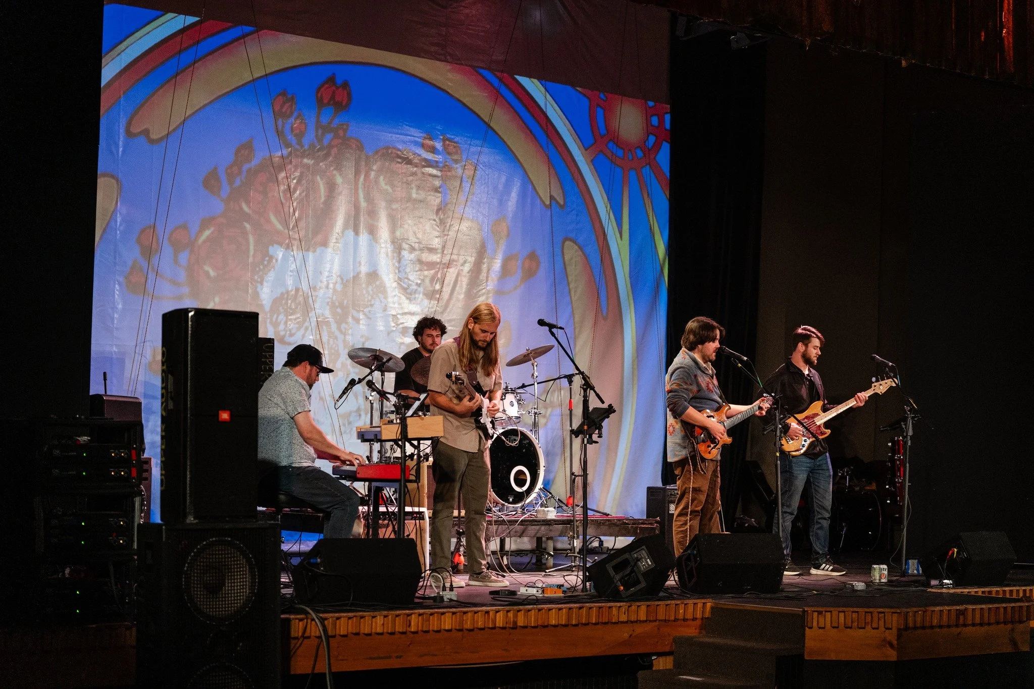 A band performing on stage with five musicians playing instruments including a keyboard, drums, bass, and two guitarists. A large colorful backdrop is projected behind them.