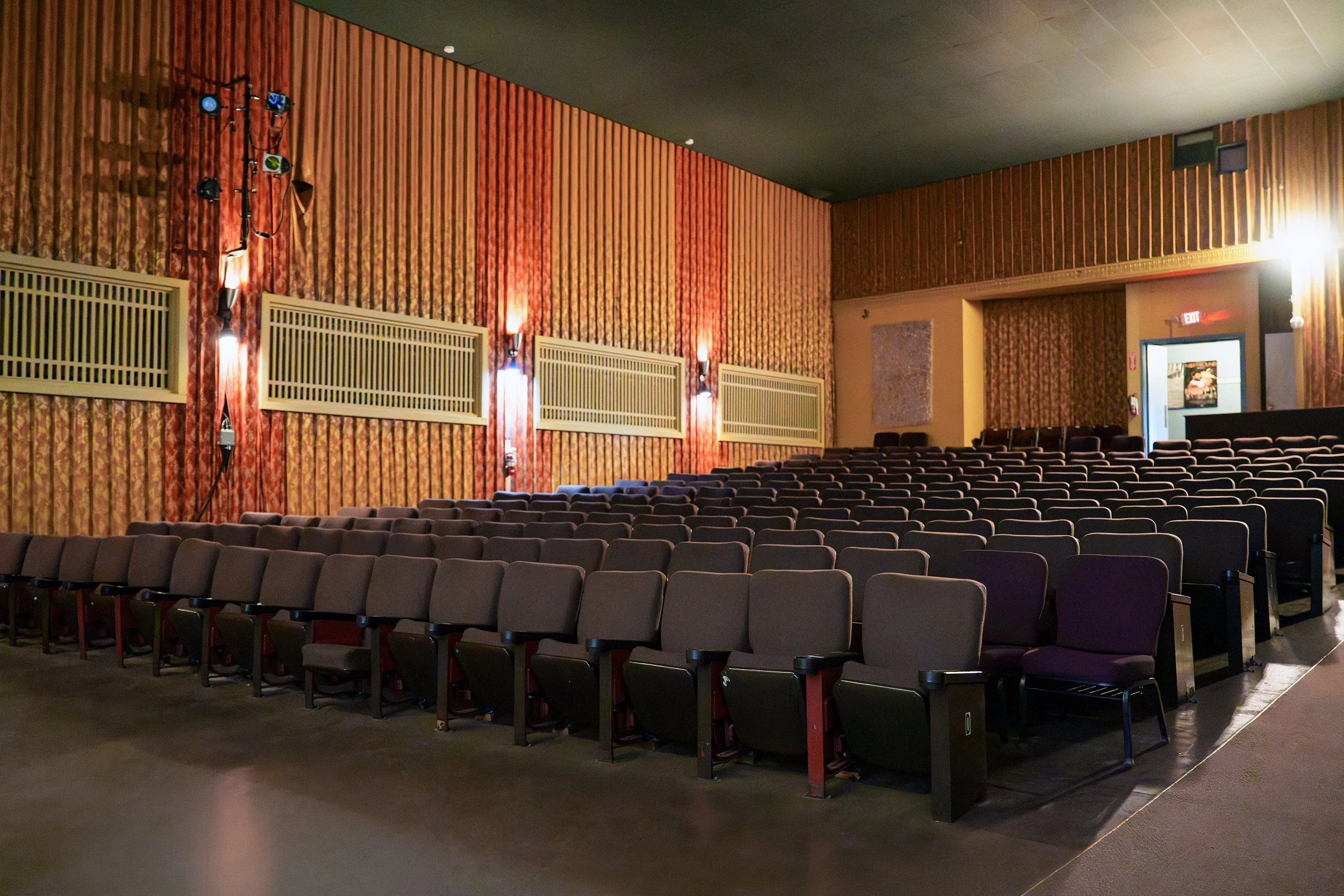 Empty theater with rows of seats, wood-paneled walls, and a door near the back wall.