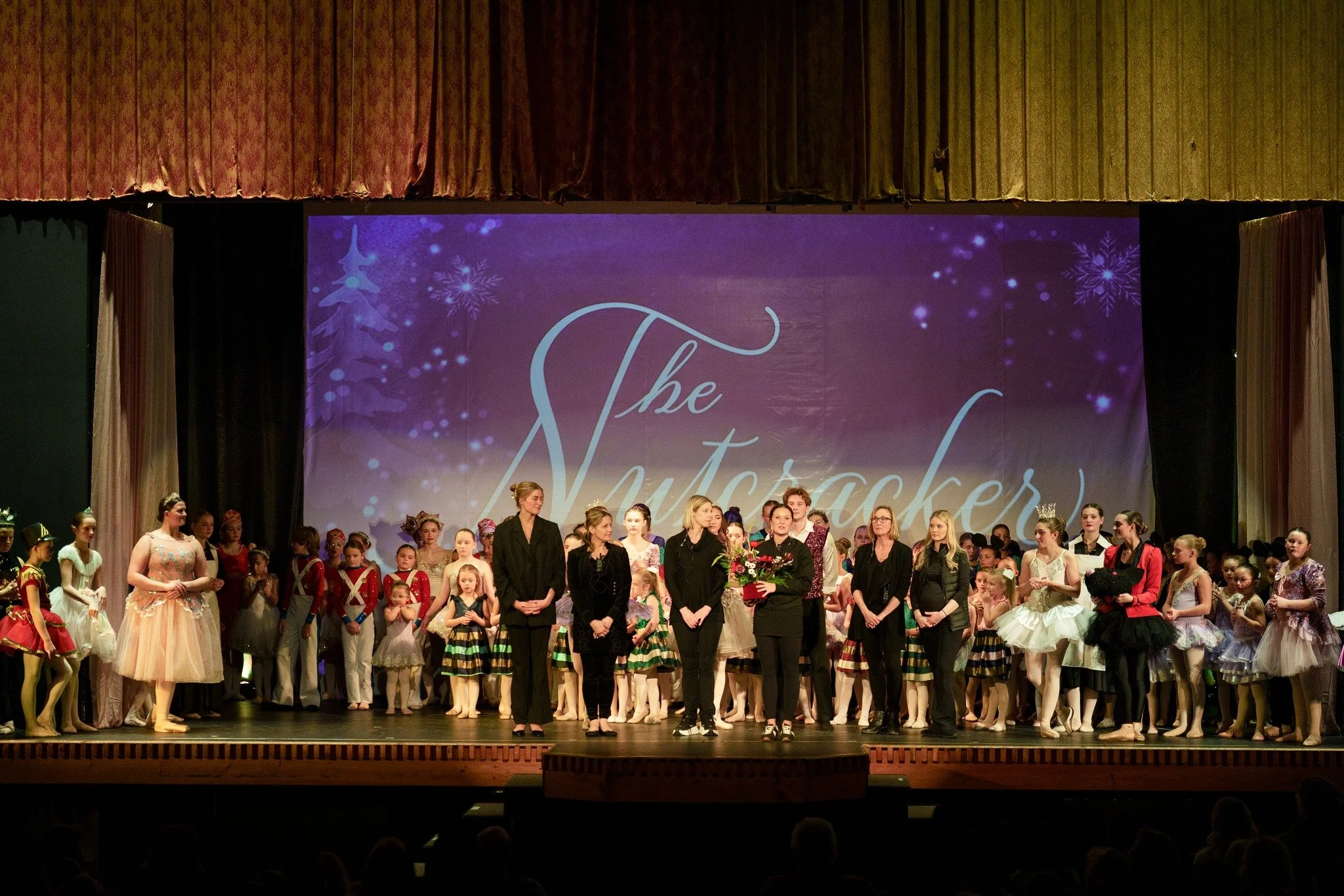 Performance on stage featuring children and adults dressed in costumes, with a backdrop saying 'The Nutcracker' and snowflake decorations.